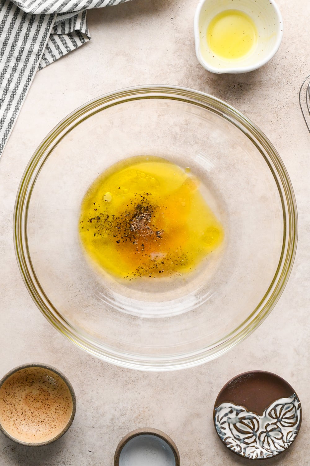 A glass bowl with olive oil, vinegar, honey, black pepper, and salt sits on a countertop, surrounded by a few small emptied prep bowls. A striped napkin is partially visible in the corner.