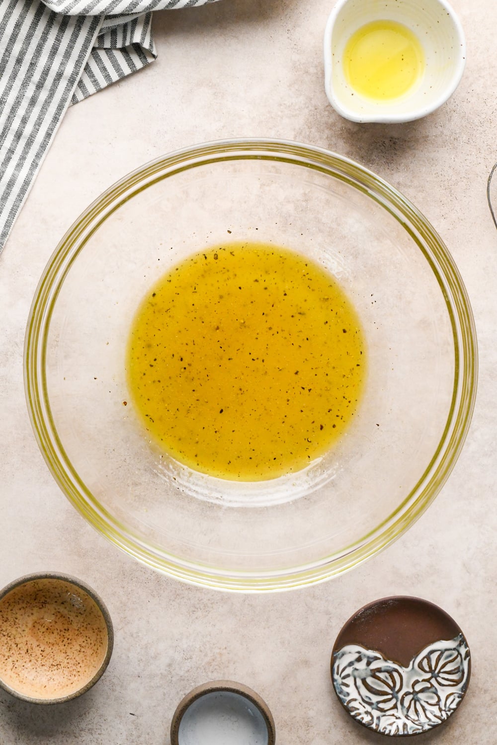 A glass bowl with yellow vinaigrette, surrounded by dishes holding oil, spices, and salt, on a light countertop with a striped cloth in the corner.