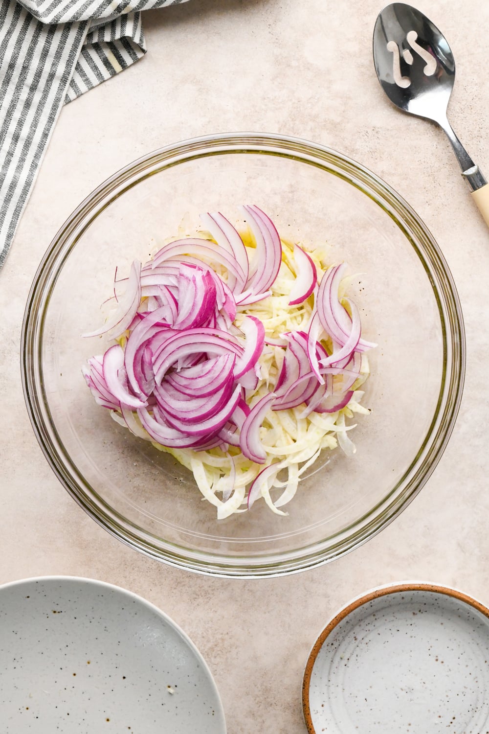A glass bowl filled with thinly sliced red onion and thinly sliced fennel, placed on a light countertop beside a striped towel, a spoon, and two empty plates.