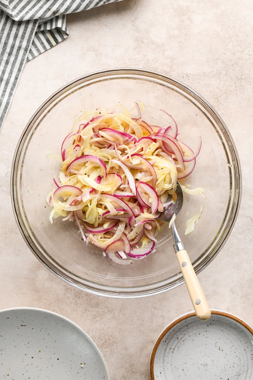 A glass bowl filled with thinly sliced red onion and thinly sliced fennel after being tossed into and marinated in the vinaigrette, placed on a light countertop beside a striped towel, a spoon, and two empty plates.