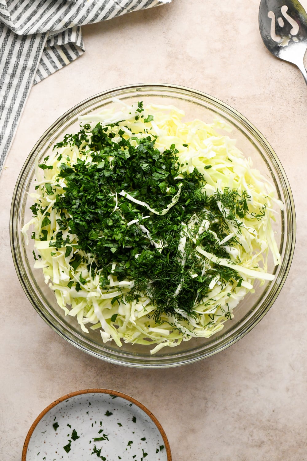 A glass bowl filled with shredded cabbage and chopped fresh herbs, including parsley and dill, sits on a light countertop next to a striped towel and a slotted spoon.