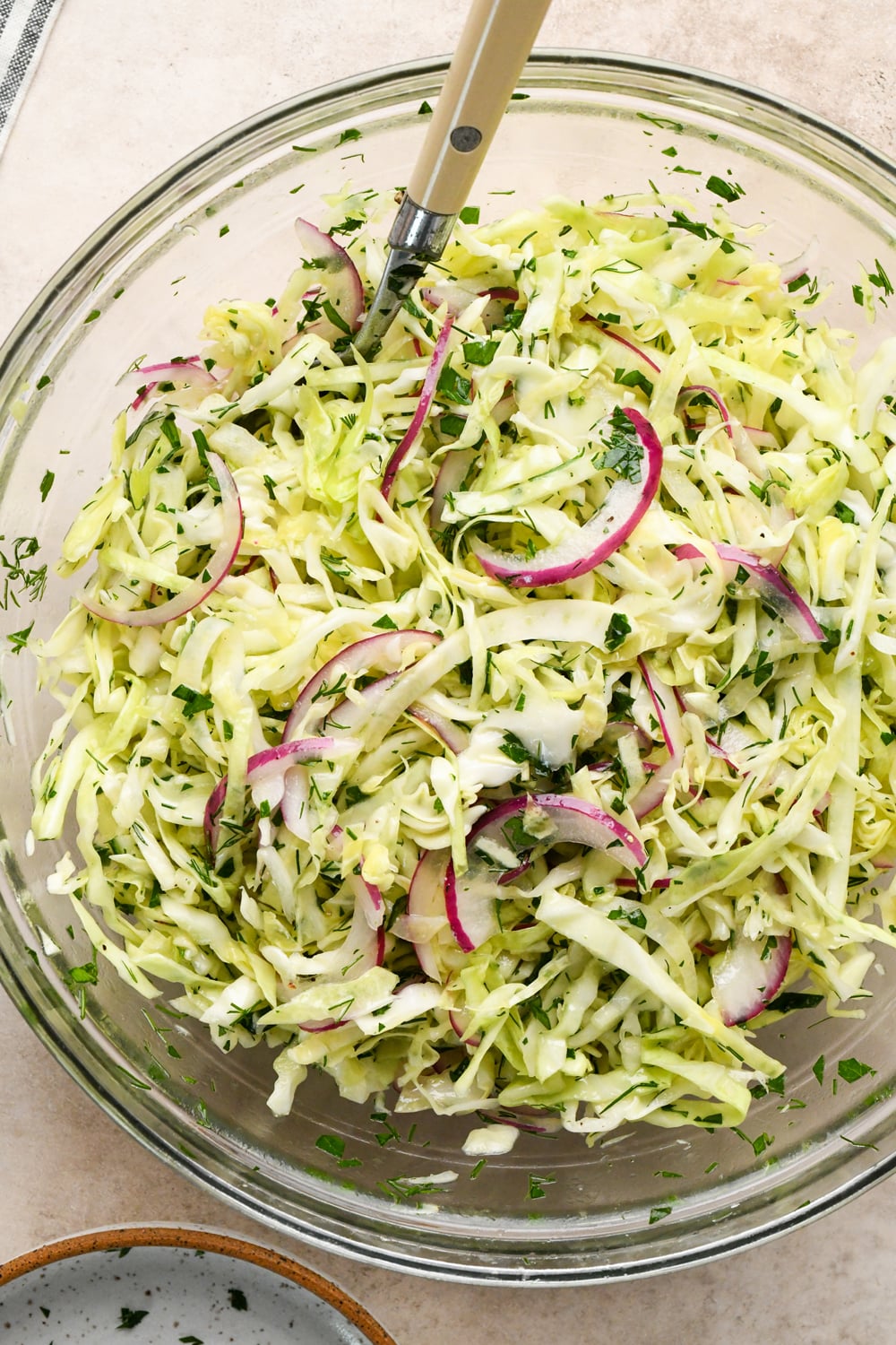 Glass bowl filled with shredded cabbage, sliced red onions, and chopped herbs, with a serving utensil resting inside the bowl. The slaw appears fresh and lightly dressed.