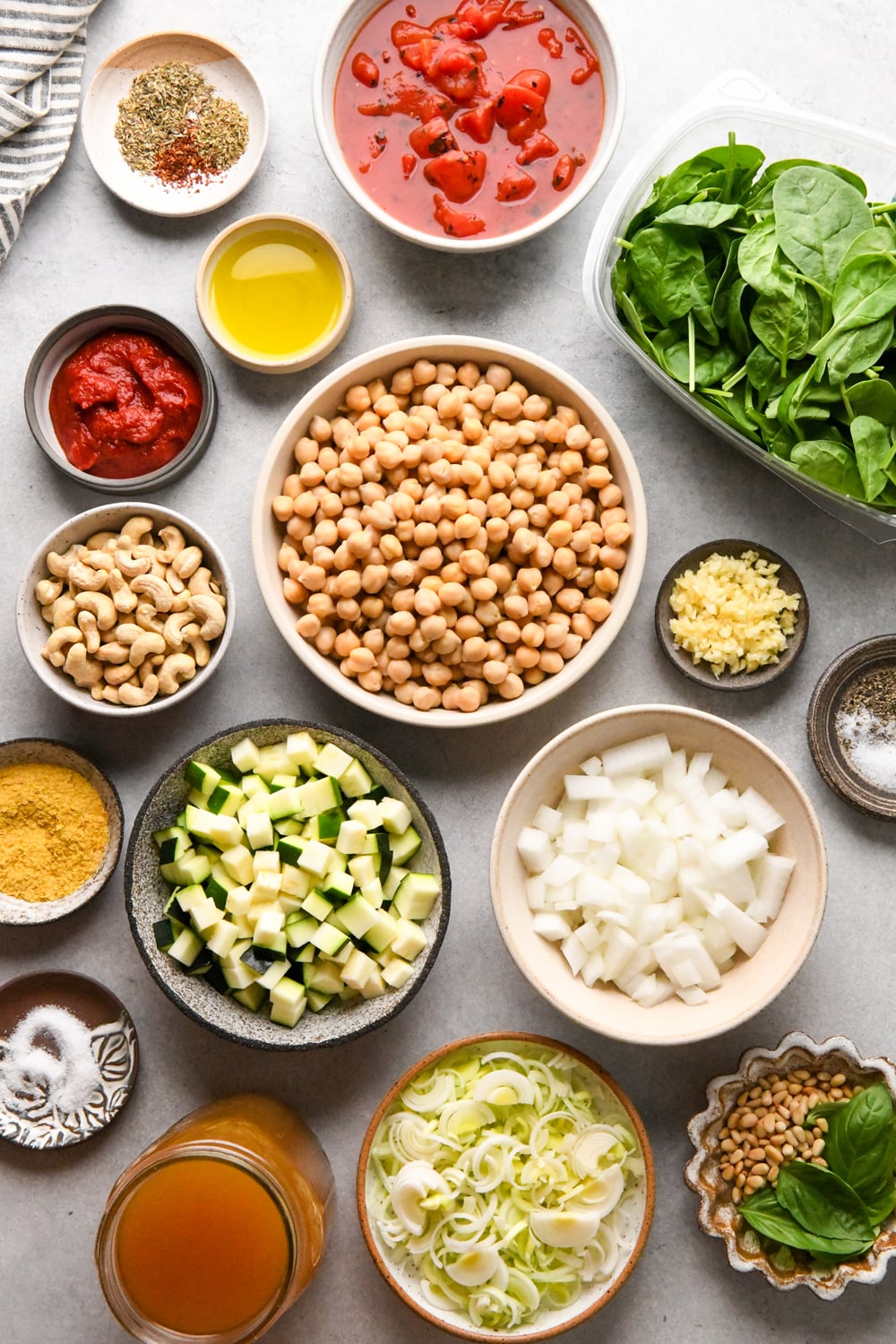 Assorted ingredients in bowls on a white surface, including chickpeas, chopped onions, zucchini, leeks, cashews, spinach, canned tomatoes, tomato paste, spices, olive oil, garlic, vegetable broth, and nutritional yeast.