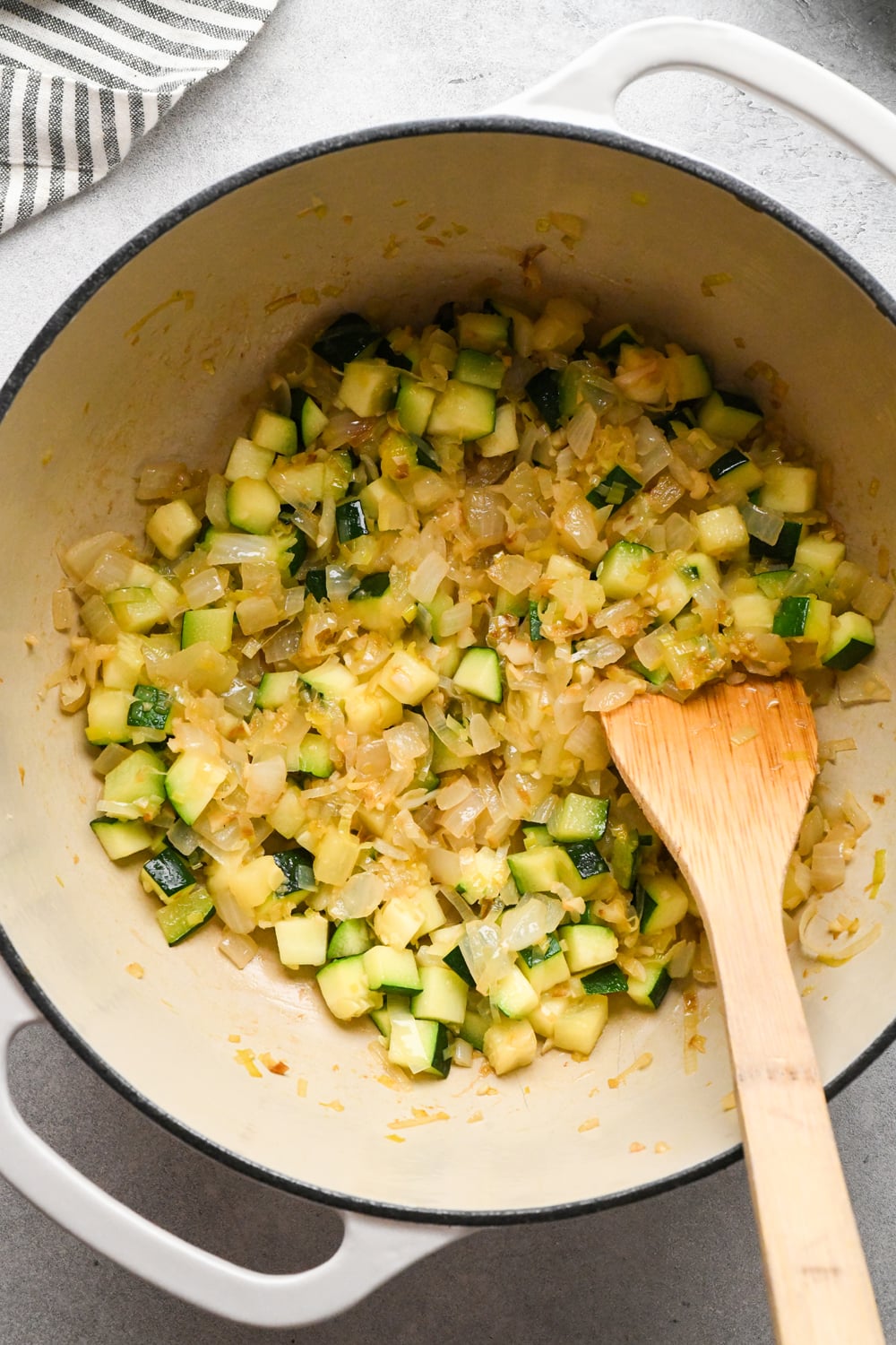 Diced zucchini in a large white pot after sautéing with the sautéed aromatics, with a wooden spoon for stirring. A striped towel is partially visible in the corner.