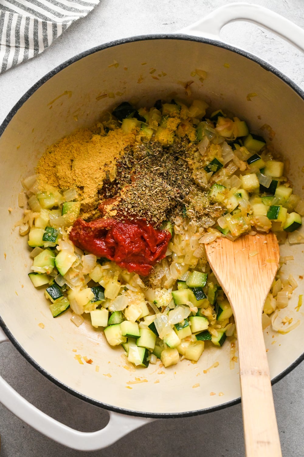 Tomato paste, nutritional yeast, Italian seasoning, and dried oregano added to the pot with the sautéed diced zucchini and aromatics, with a wooden spoon for stirring. A striped towel is partially visible in the corner.