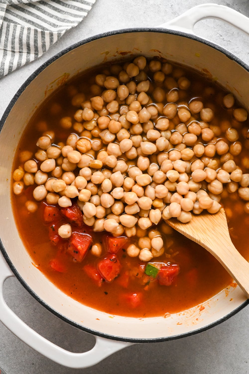 A white pot filled with chickpeas, diced tomatoes, and broth being stirred with a wooden spoon, sitting on a light countertop near a striped towel.