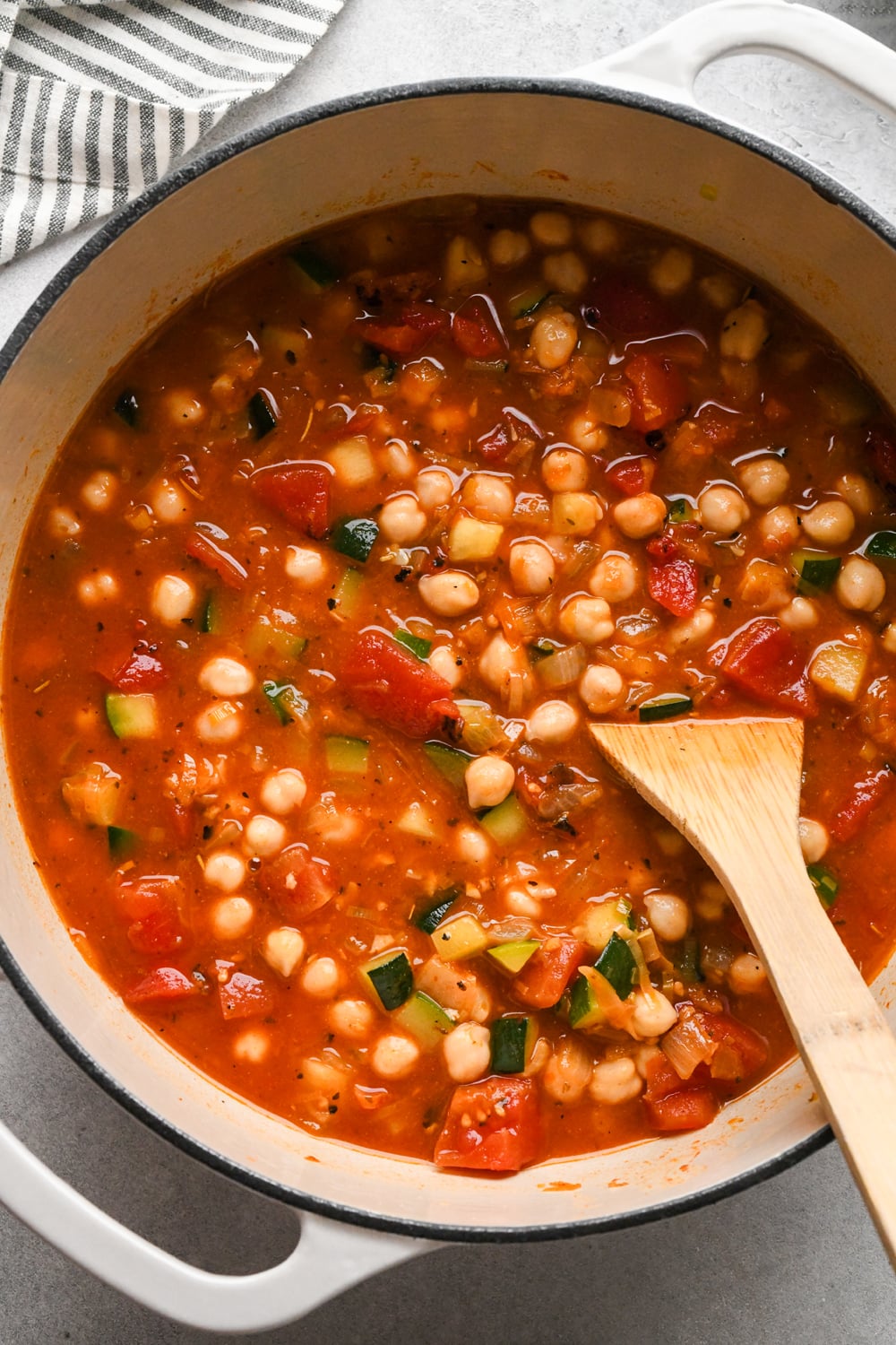 A pot of vegetable and chickpea soup with tomatoes, zucchini, and spices is being stirred with a wooden spoon. A striped cloth is visible in the corner.