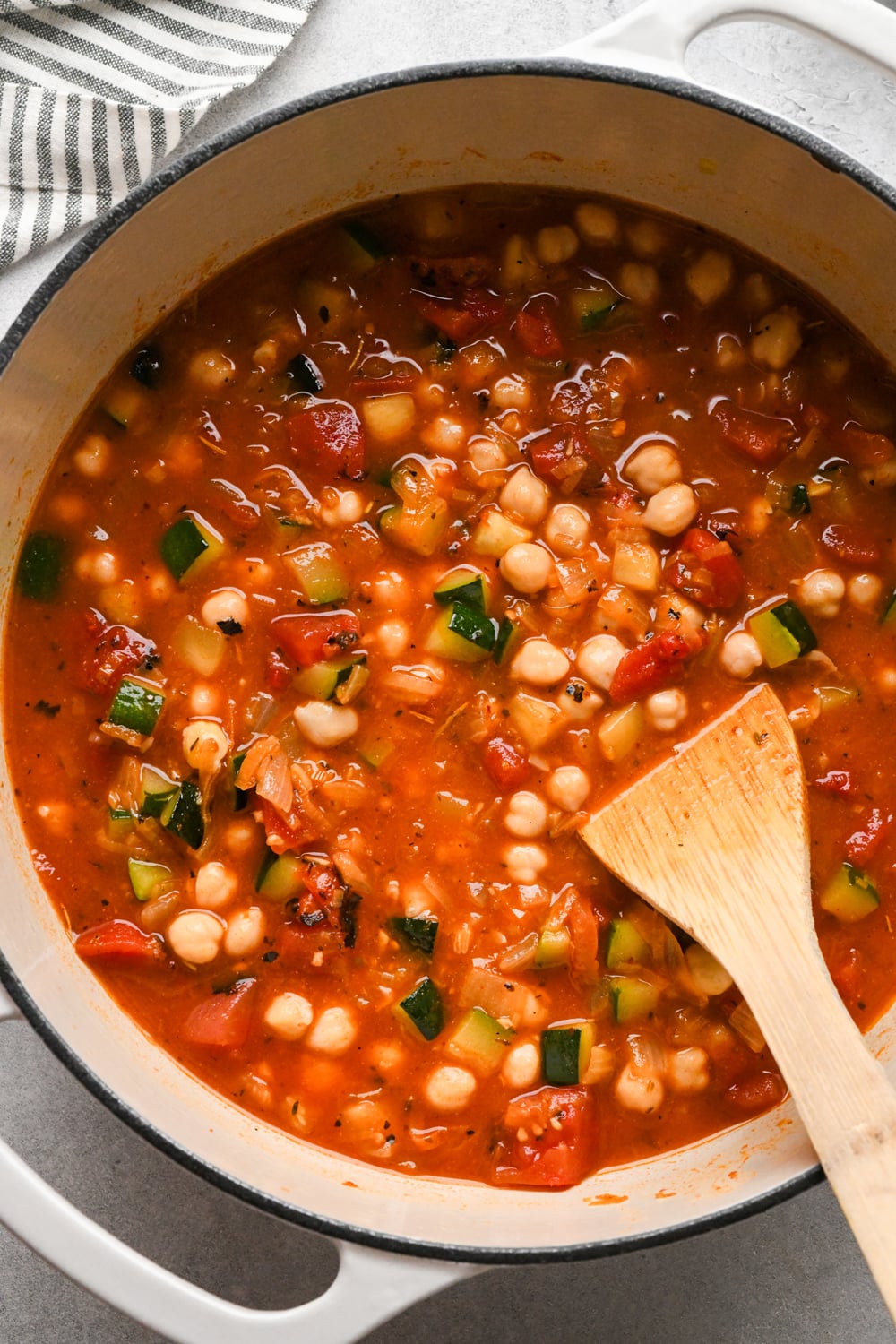A pot of vegetable and chickpea soup with tomatoes, zucchini, and spices is being stirred with a wooden spoon after having simmered for a short while to reduce. A striped cloth is visible in the corner.