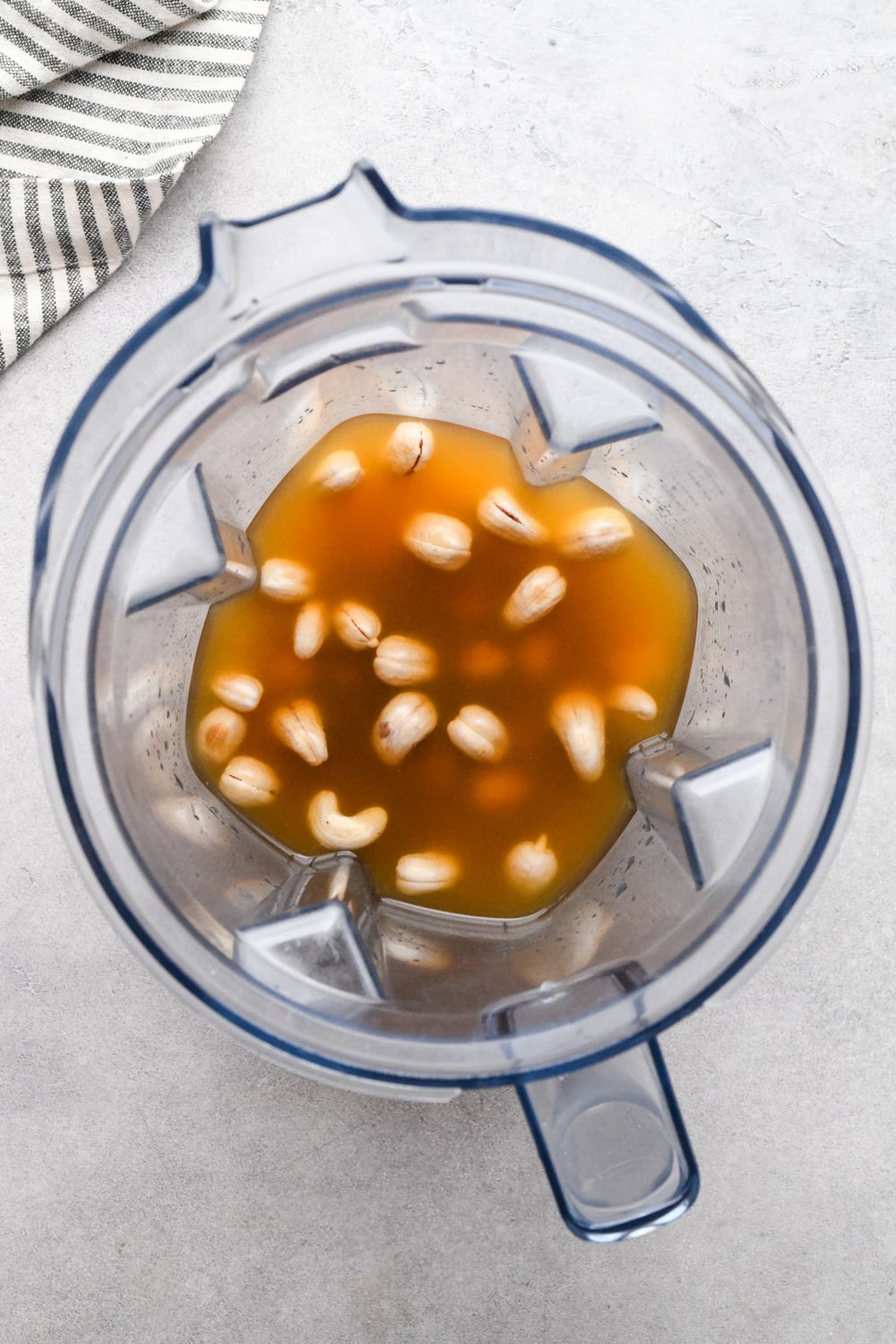 Top view of a blender with vegetable broth and soaked raw cashews, placed on a light countertop next to a striped cloth.