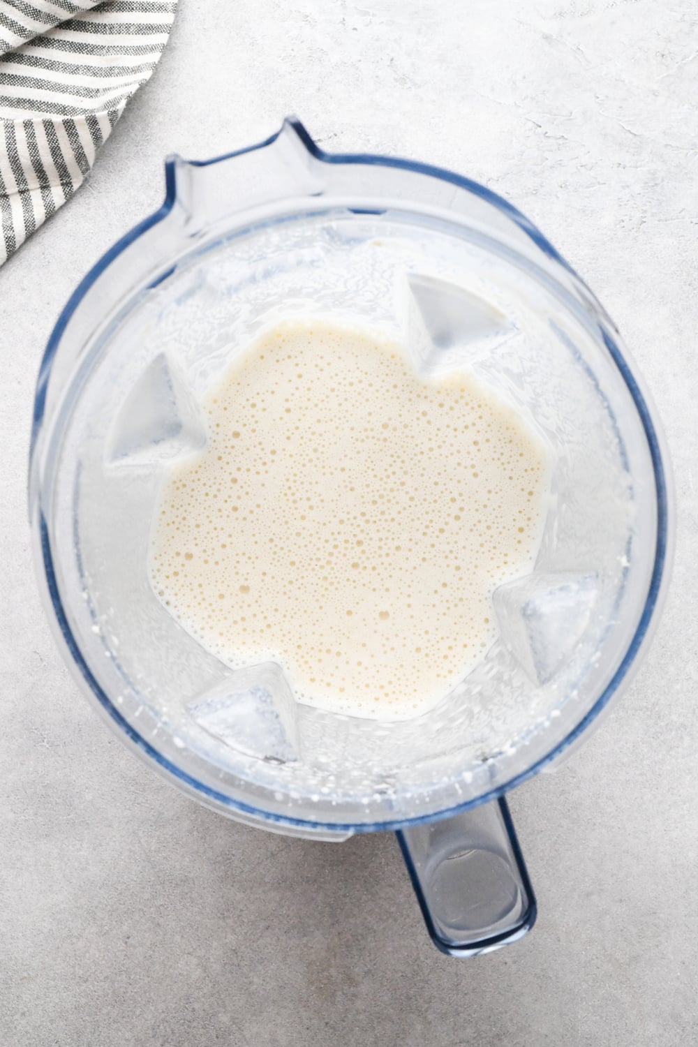 Top view of a blender with vegetable broth and soaked raw cashews after blending into a creamy mixture, placed on a light countertop next to a striped cloth.