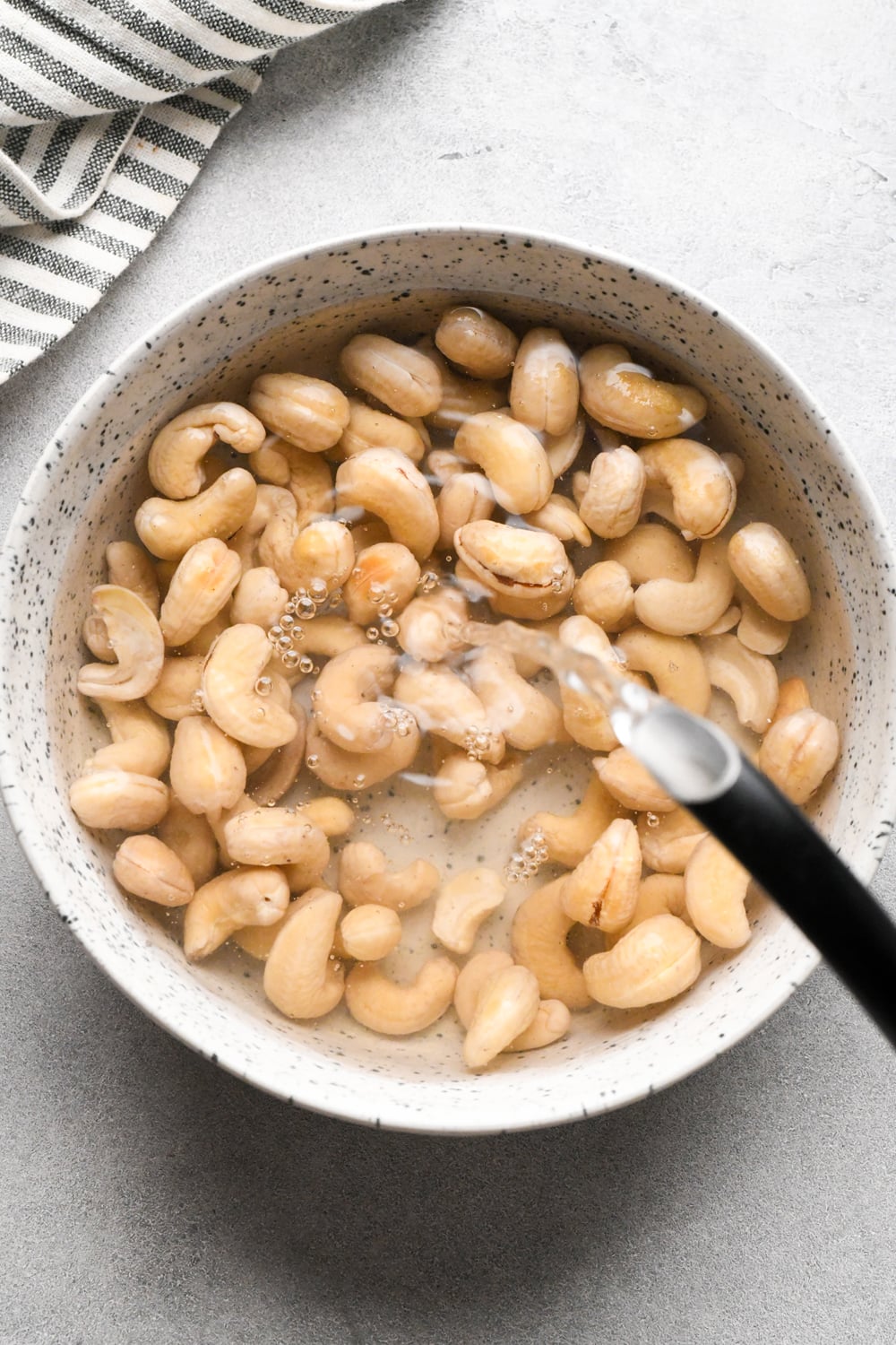 A bowl of raw cashews is being filled with water from a black tea kettle spout, preparing them for soaking. A striped towel is partially visible in the corner.