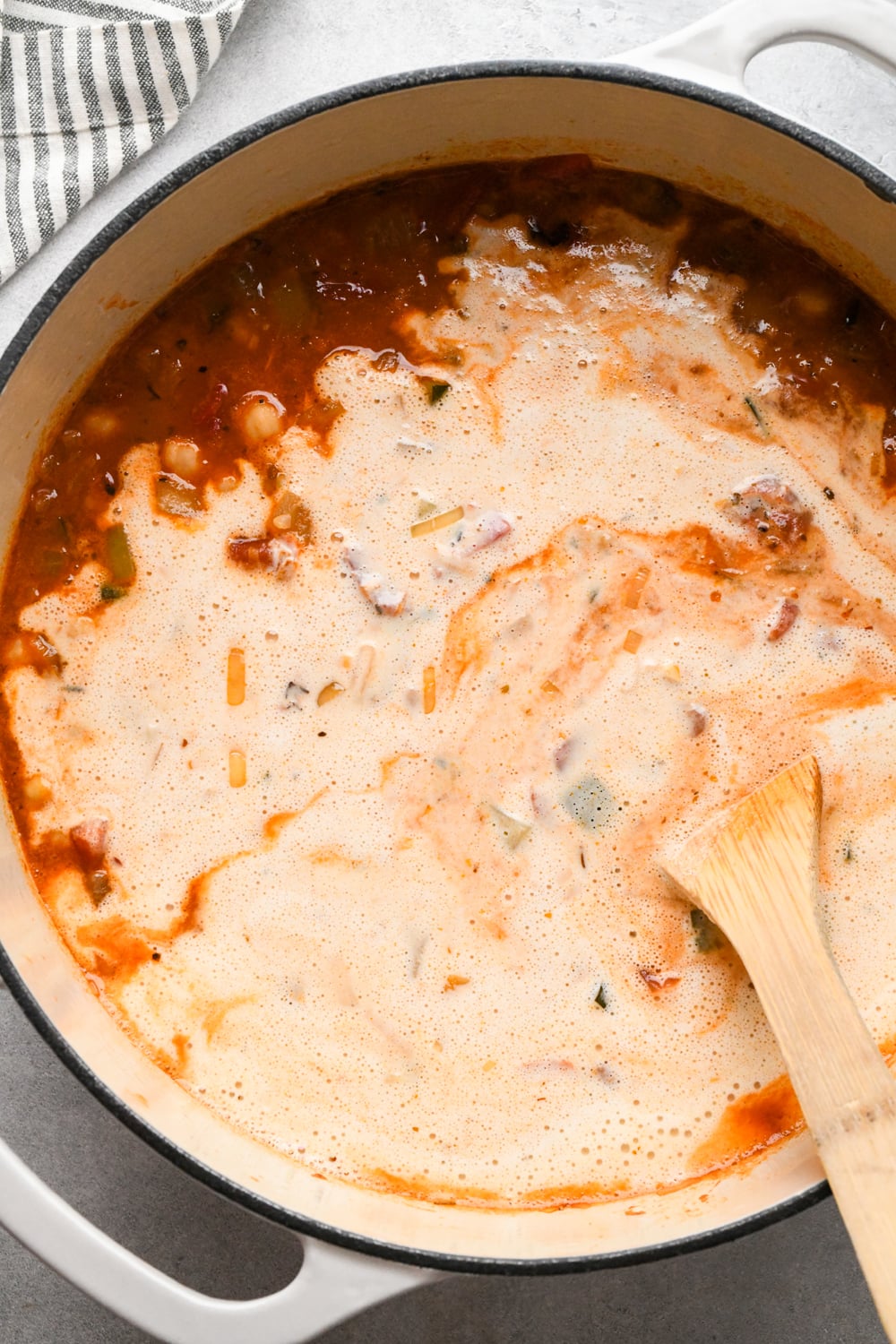 A pot of vegetable and chickpea soup with tomatoes, zucchini, and spices is being stirred with a wooden spoon, white cashew cream poured into the pot. A striped cloth is visible in the corner.