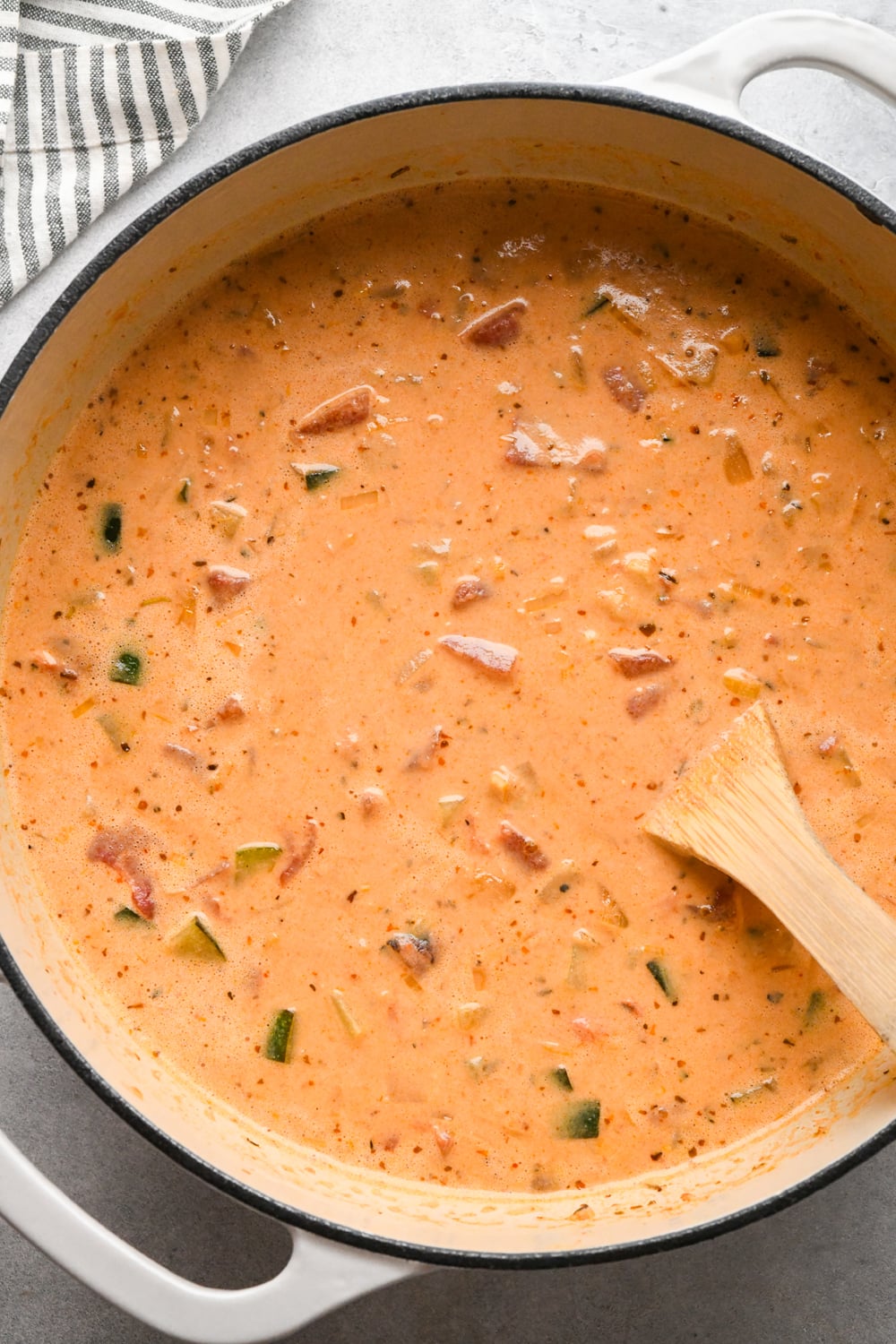 A creamy, orange-colored soup with visible chunks of vegetables and chickpeas simmers in a large pot. A wooden spoon rests inside, and a striped towel is nearby on the gray surface.