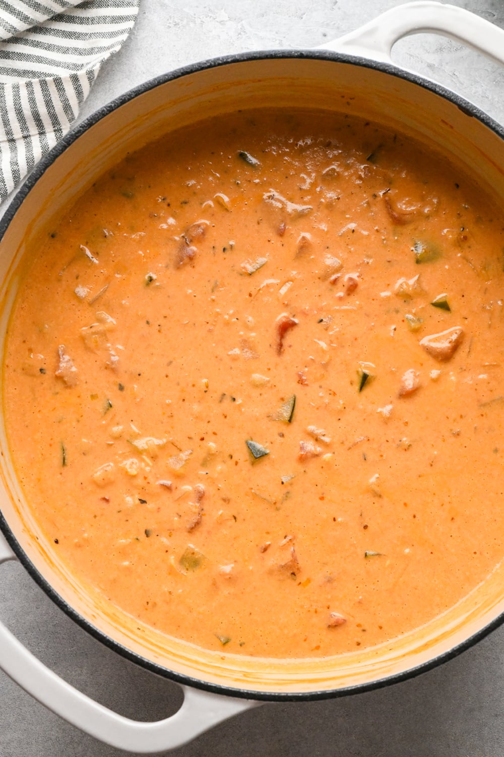 A creamy, orange-colored soup with visible chunks of vegetables and chickpeas after simmering to reduce slightly in a large pot. A wooden spoon rests inside, and a striped towel is nearby on the gray surface.