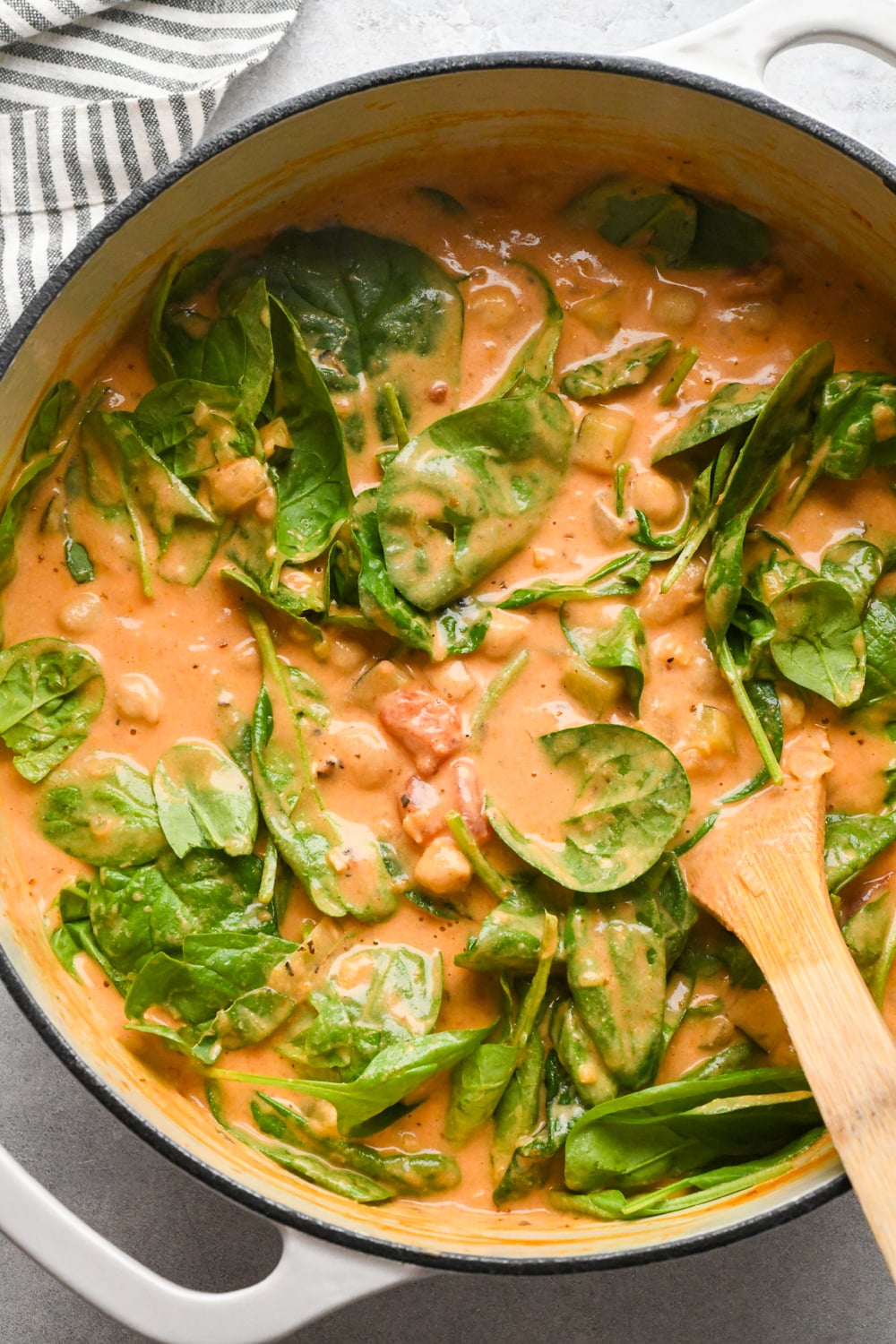 A creamy tomato-based stew with fresh spinach leaves and chunks of vegetables in a white pot with a wooden spoon, next to a striped cloth napkin.