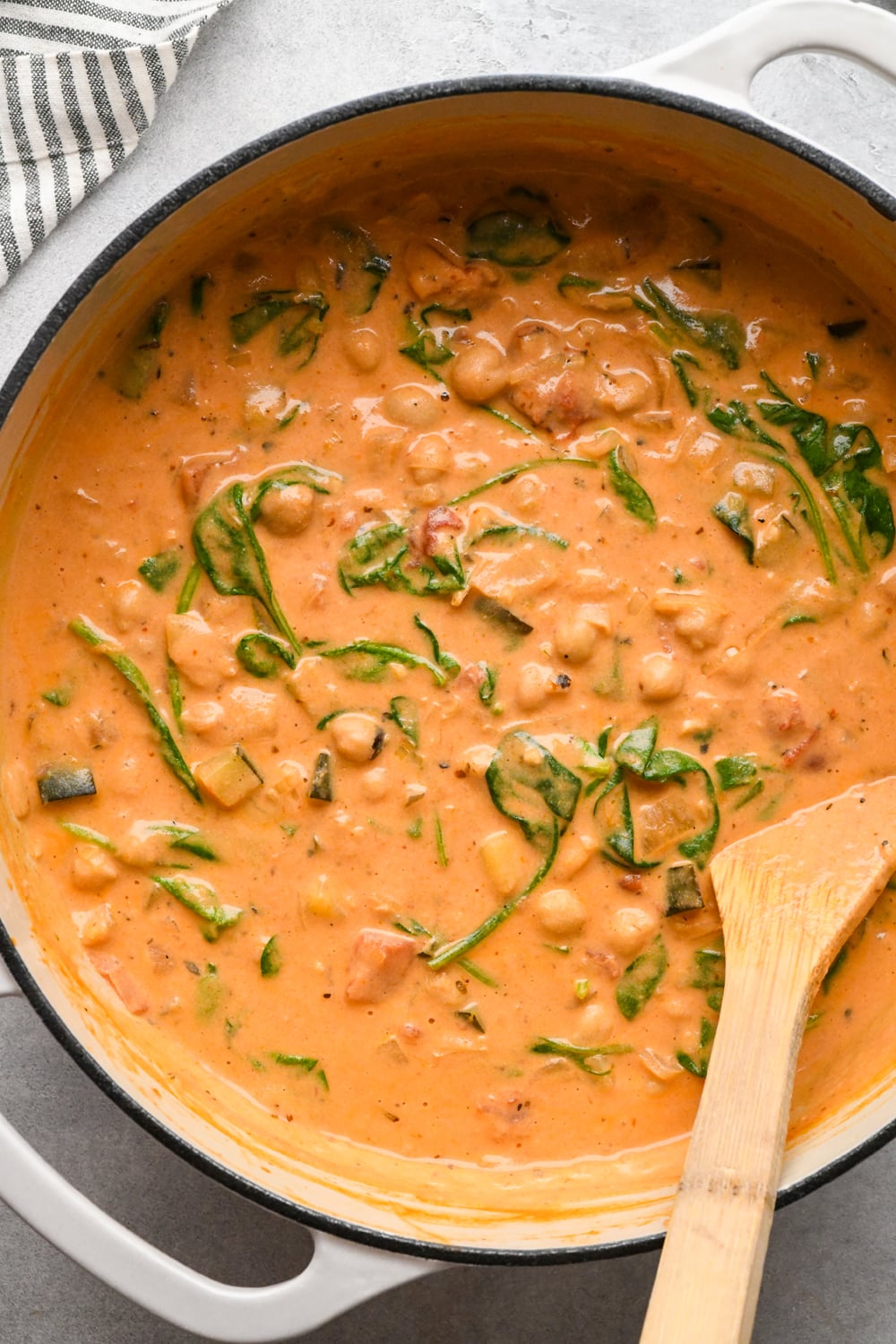 A pot filled with creamy tomato-based soup with chickpeas, spinach, and diced vegetables. A wooden spoon rests on the edge of the pot.