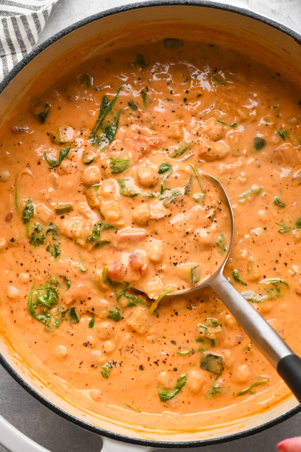 A pot of creamy tomato-based chickpea and veggie soup, with a ladle lifting out a portion of the dairy-free soup, topped with black pepper.