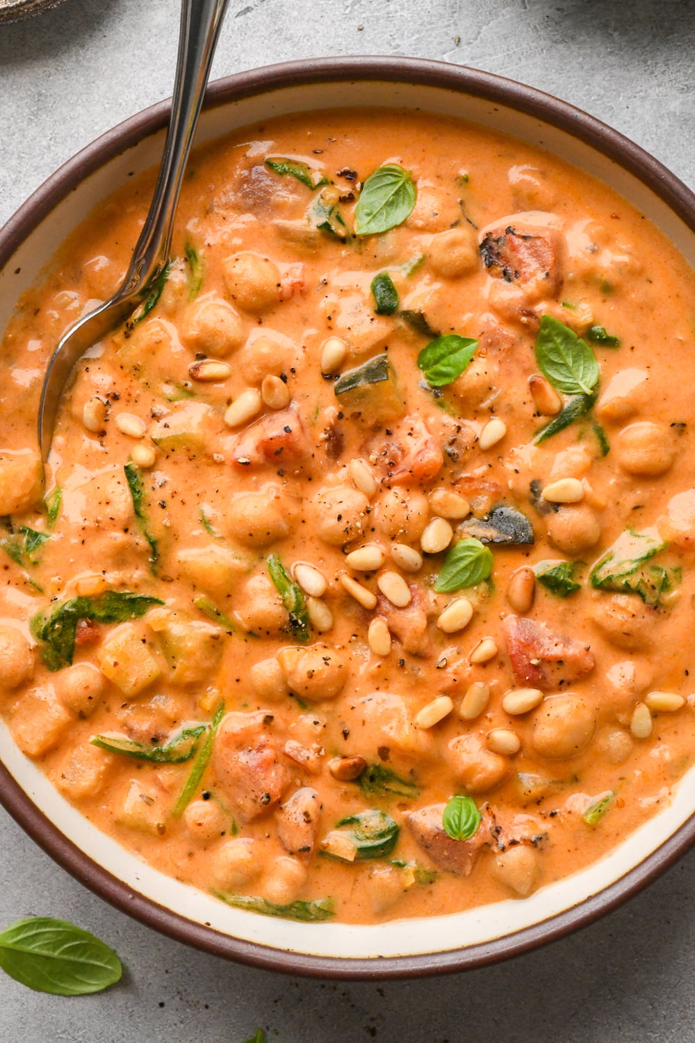 A bowl of creamy tomato-based soup with chickpeas, spinach, tomatoes, pine nuts, and fresh basil, with a spoon resting in the bowl.