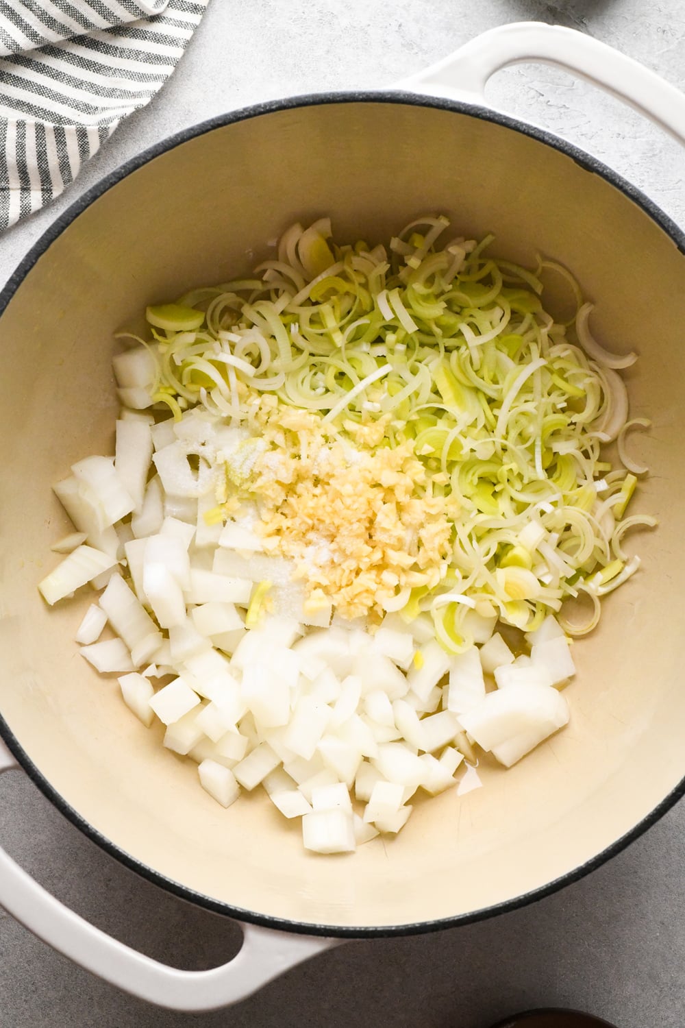 Chopped onions, leeks, and minced garlic are in a large white pot, ready to be cooked. A striped towel is partially visible in the corner.