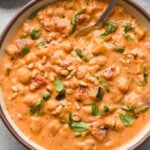 A bowl of creamy tomato chickpea soup garnished with fresh basil leaves and pine nuts, with a spoon inside. The soup has visible chickpeas,zucchini, spinach, and fire-roasted canned tomato pieces.