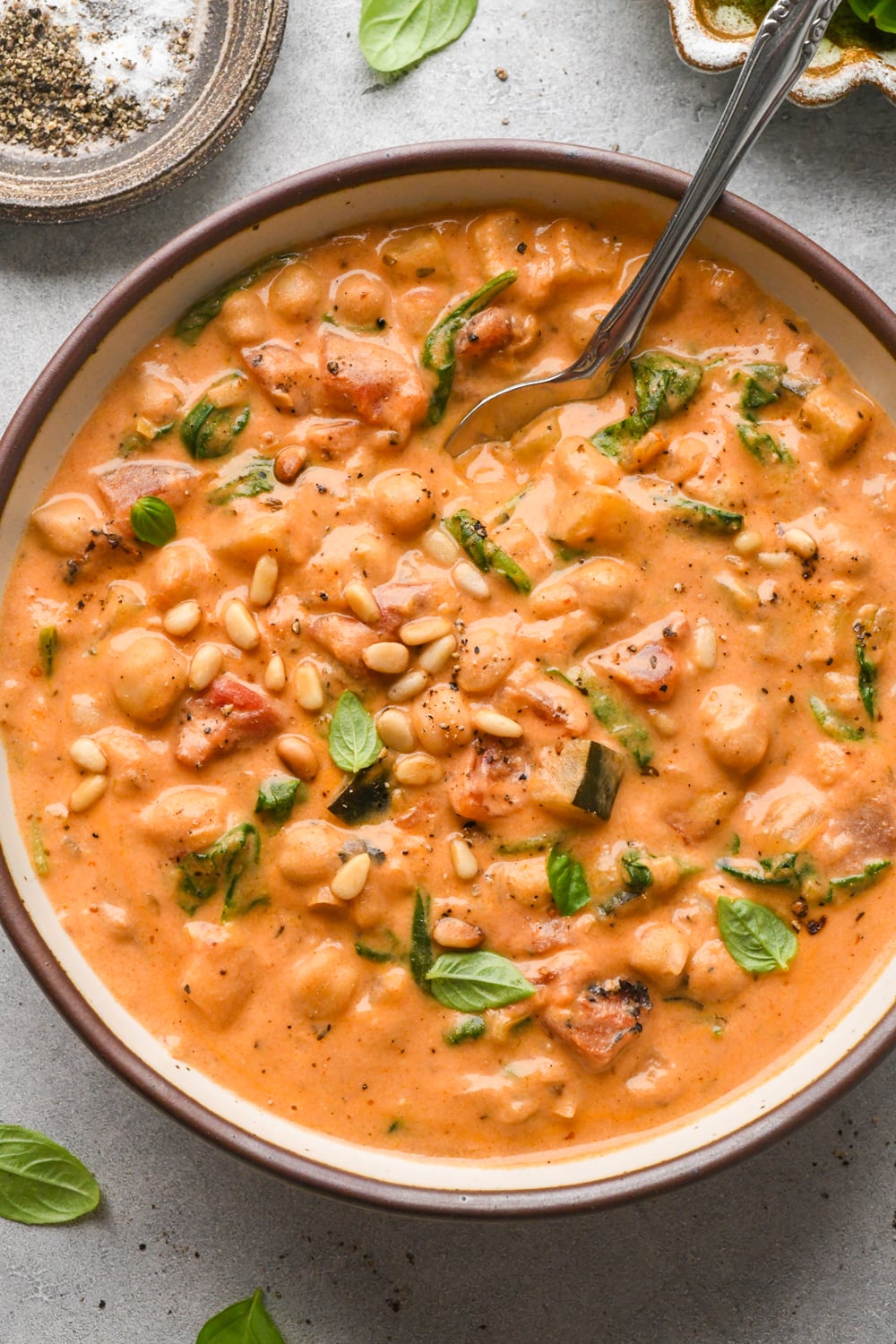 A bowl of creamy tomato chickpea soup garnished with fresh basil leaves and pine nuts, with a spoon inside. The soup has visible chickpeas,zucchini, spinach, and fire-roasted canned tomato pieces.