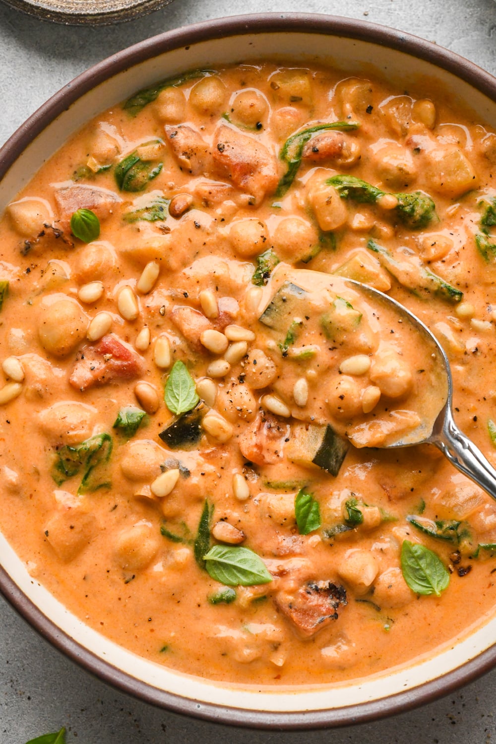 A bowl of creamy tomato chickpea soup with spinach, garnished with pine nuts and fresh basil leaves, with a spoon lifting out a bite of the soup.