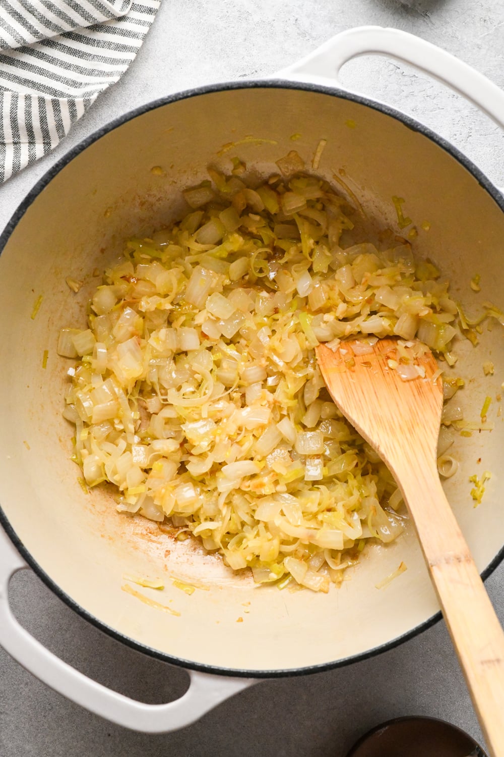Chopped onions and leeks sautéing in a white pot, stirred with a wooden spoon. The mixture is slightly browned, with a striped cloth visible in the corner.