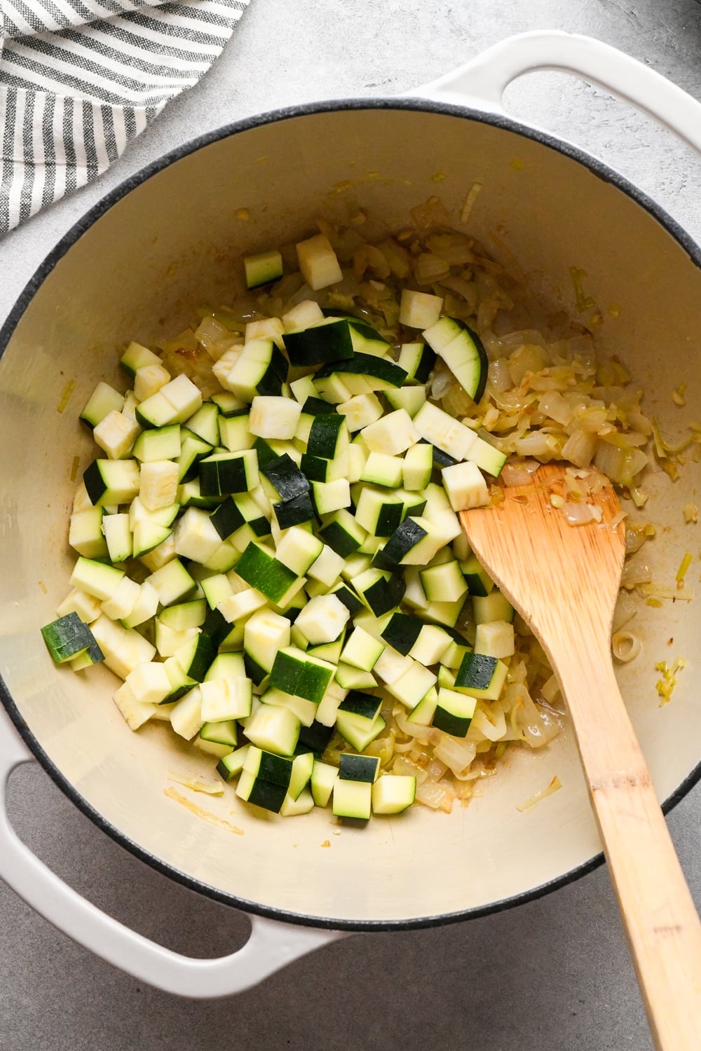 Raw, diced zucchini added to the pot with the sautéed aromatics, with a wooden spoon for stirring. A striped towel is partially visible in the corner.
