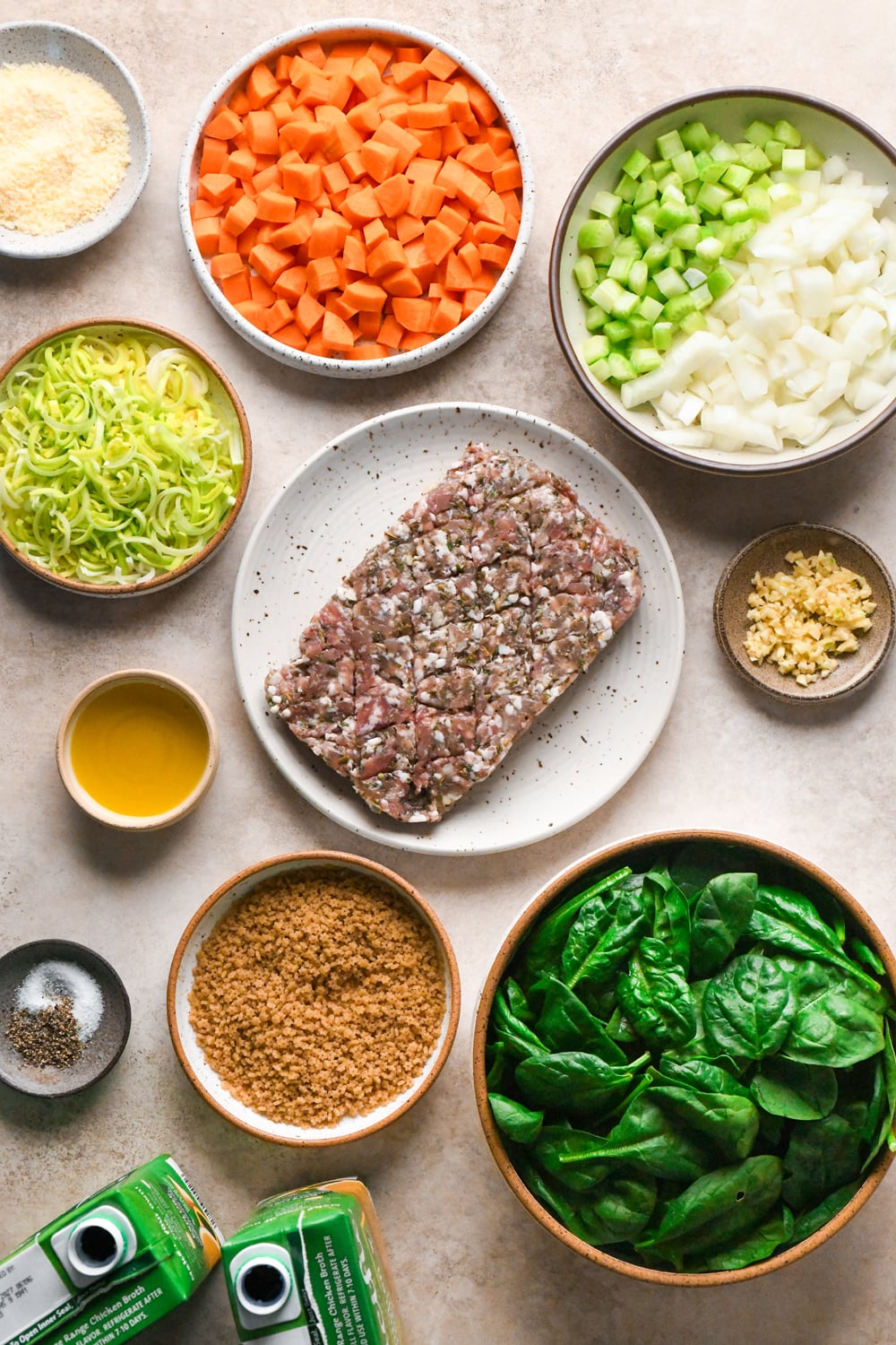 Top-down view of ingredients for making Italian sausage soup: diced carrots, chopped celery and onions, sliced leeks, ground sausage, minced garlic, spinach, gluten-free stelline pasta, chicken broth, olive oil, and seasonings.