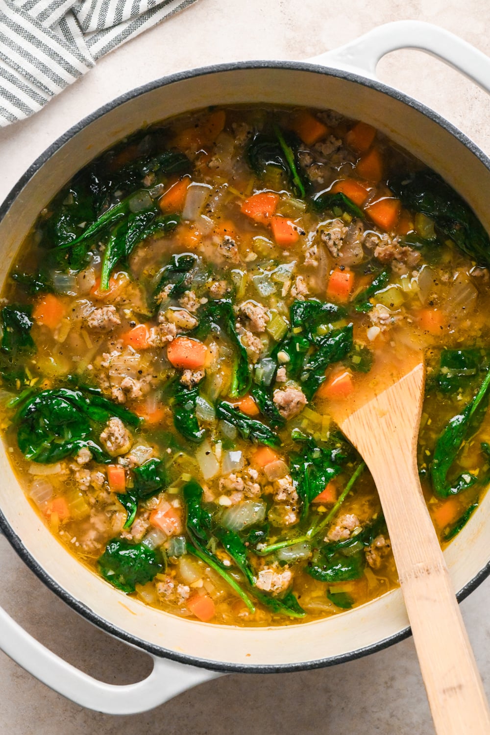 A white pot filled with soup containing ground Italian sausage, chopped carrots, celery, onion, and spinach, with a wooden spoon resting inside.