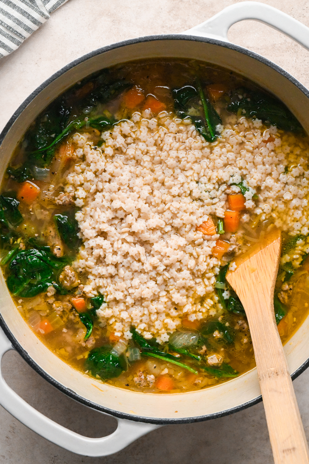 A white Dutch oven filled with soup containing star-shaped pasta, spinach, carrots, and onions, with a wooden spoon resting inside.