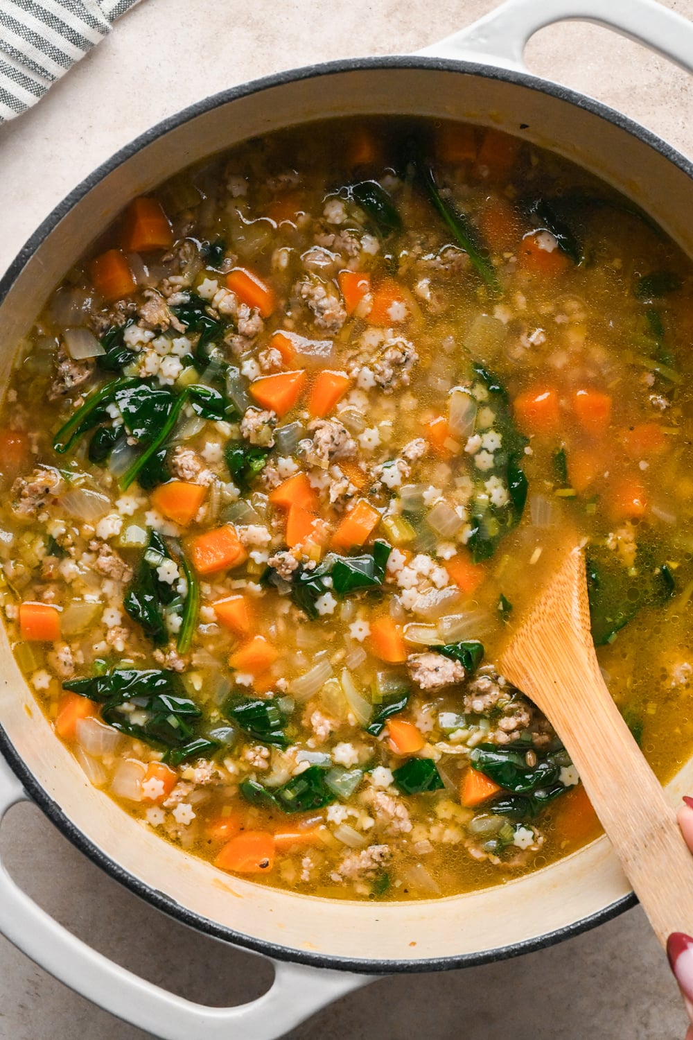 A white Dutch oven filled with soup containing star-shaped pasta, ground meat, carrots, and spinach, with a wooden spoon stirring the broth.