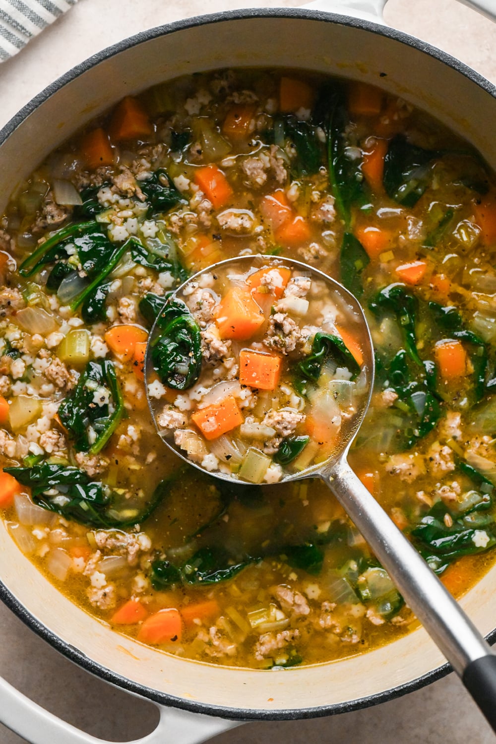 A ladle scoops vegetable and ground Italian sausage soup with spinach and pasta from a large pot.