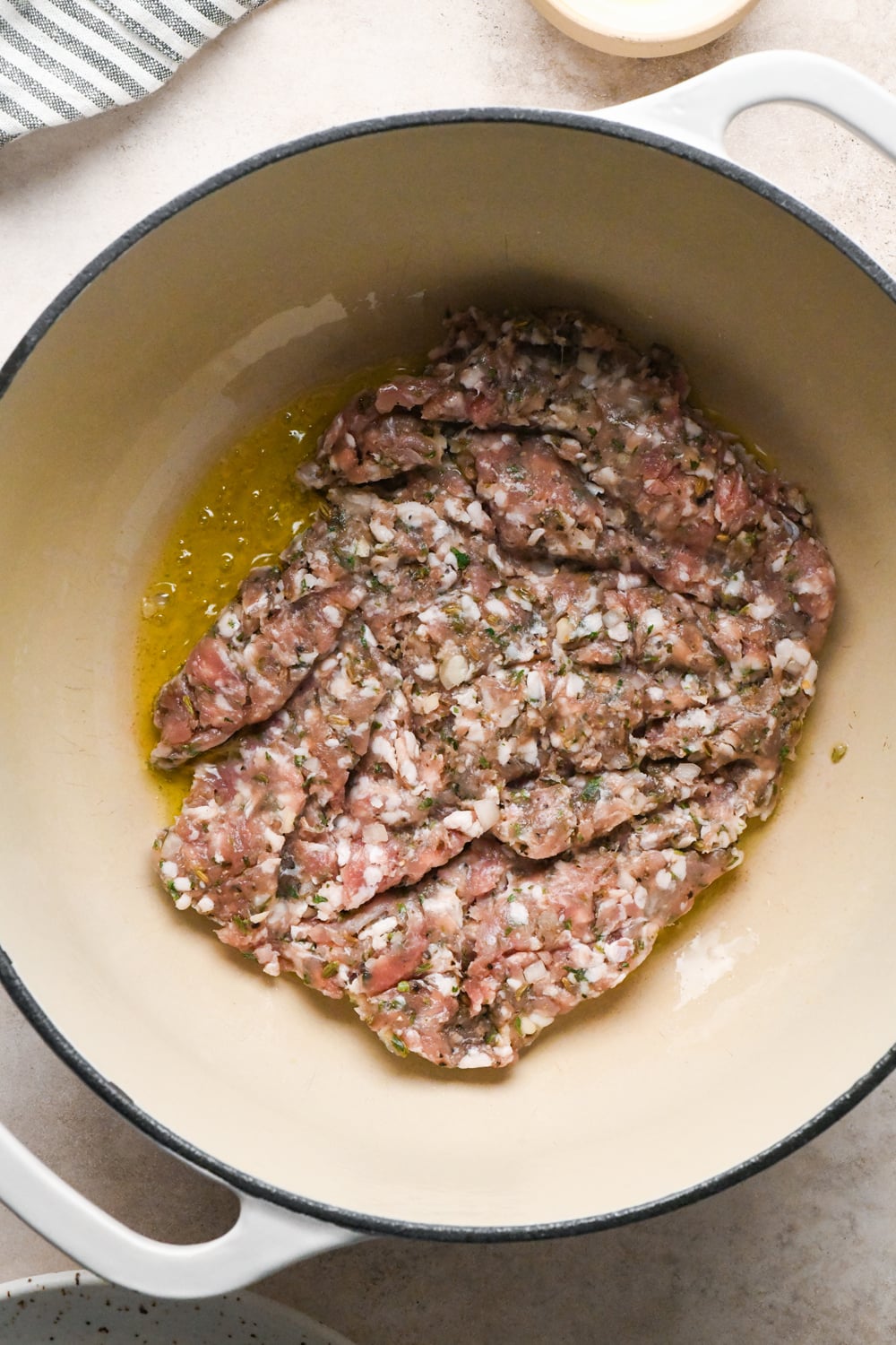 Raw seasoned ground Italian sausage pressed flat and cooking in a large white enameled Dutch oven.