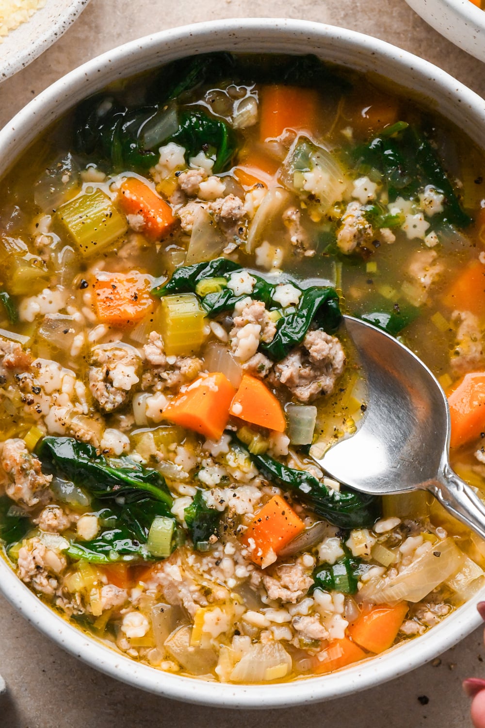 A metal spoon dipping into a bowl of soup containing ground Italian sausage, spinach, carrots, celery, and small star-shaped pasta in broth.