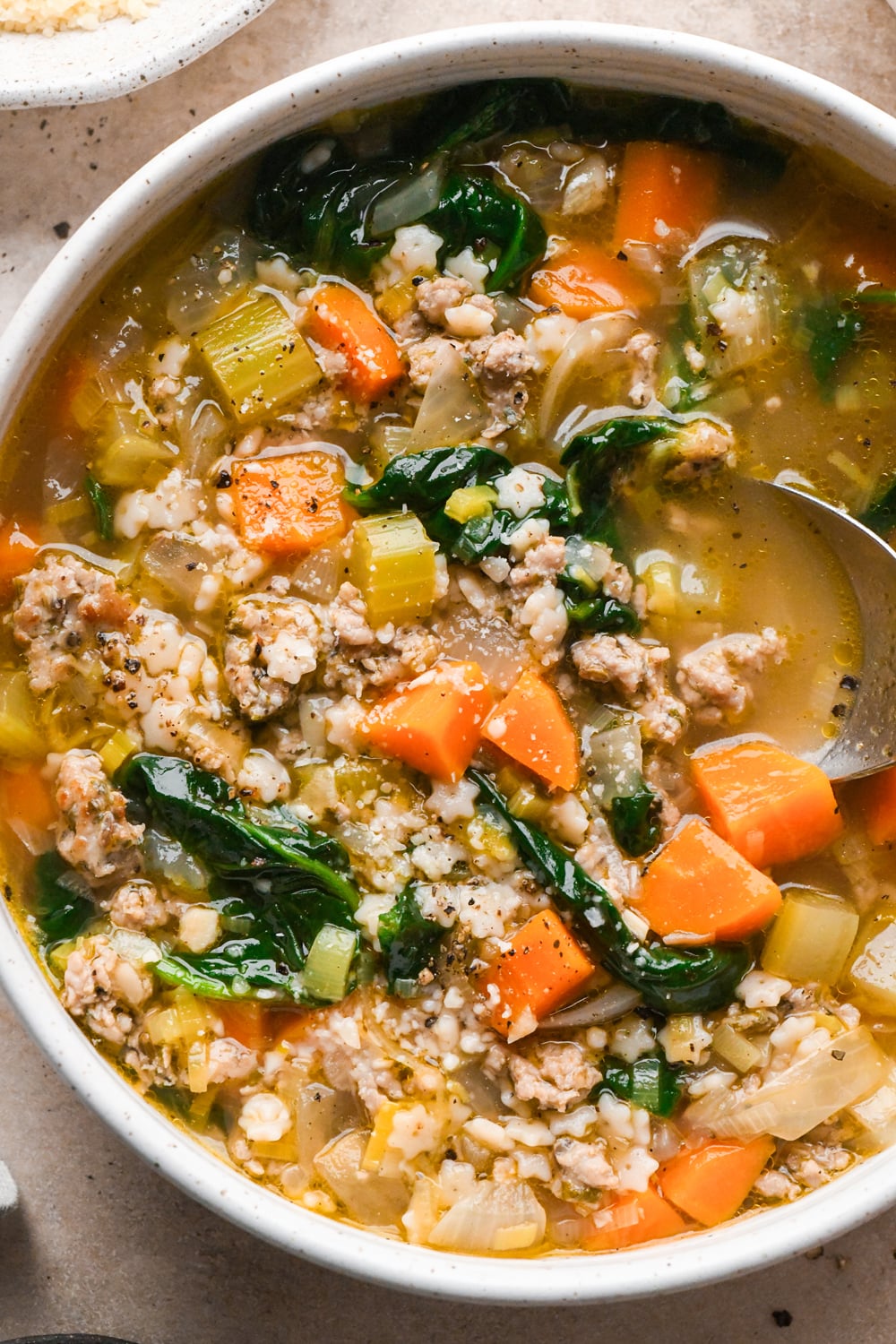 A close up image of a metal spoon dipping into a bowl of soup containing ground meat, spinach, carrots, celery, and small pasta shapes with grated parmesan in broth.