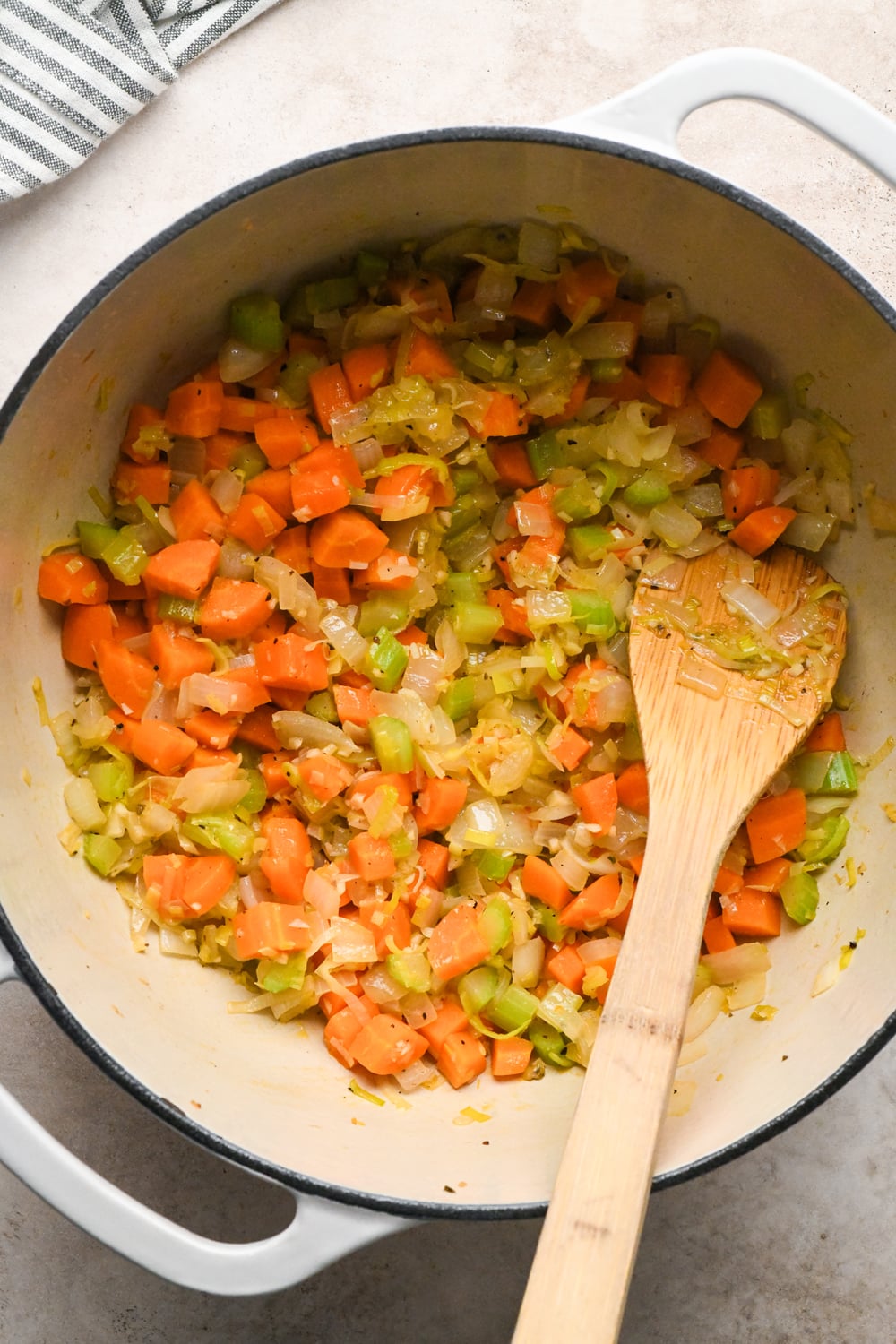 A wooden spoon stirs a mixture of sautéed diced carrots, celery, leeks, garlic, and onions in a white enameled cast iron pot.