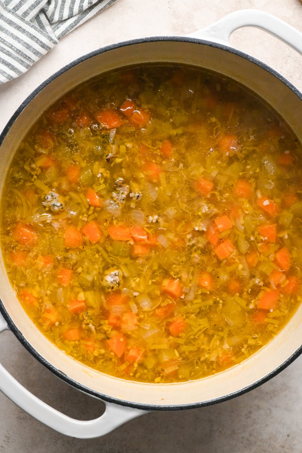 Chicken broth with other soup ingredients in a large white enameled dutch oven, after simmering so that the soup has reduced slightly and has a deeper golden hue.