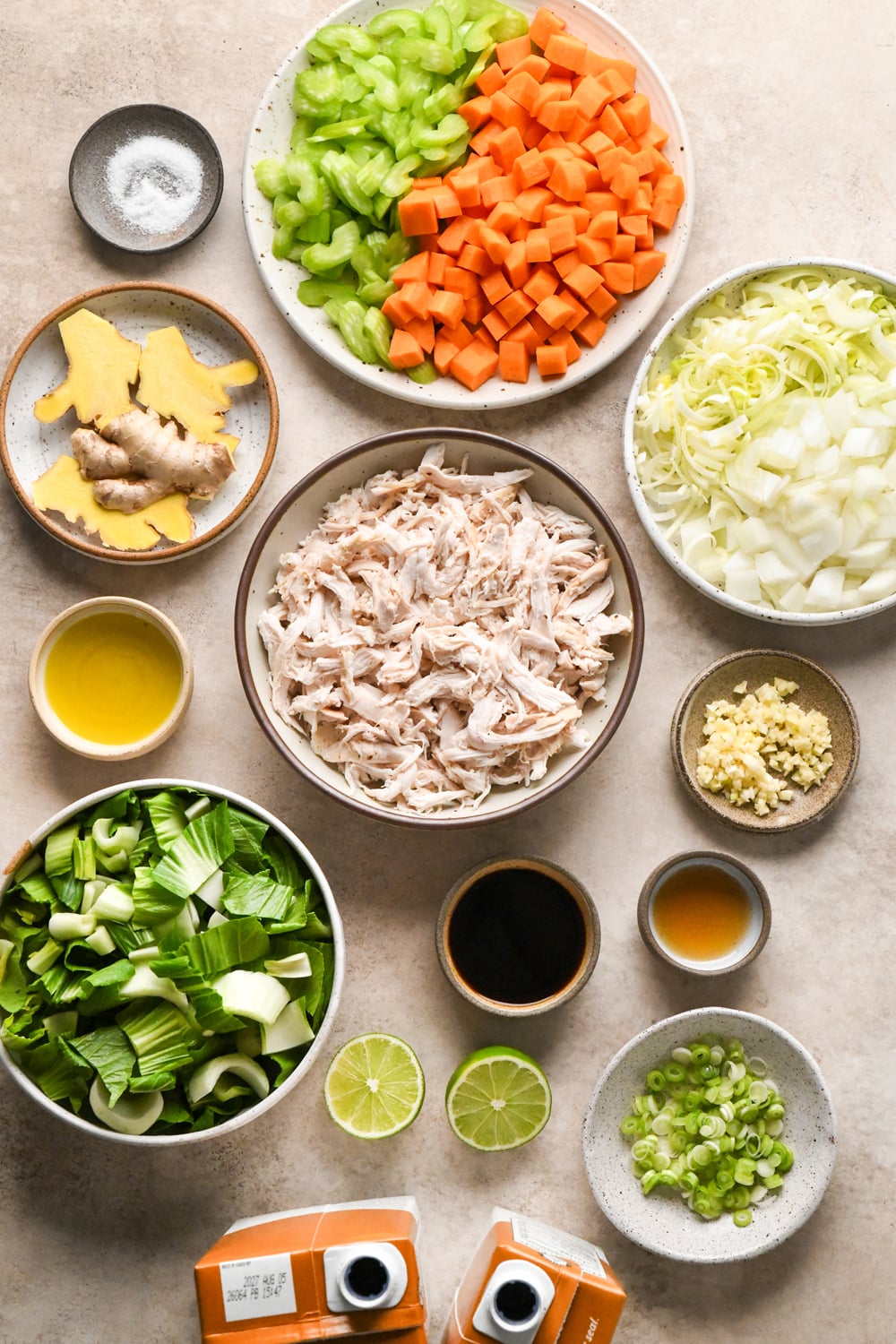 Top-down view of ingredients for chicken soup: shredded chicken, chopped carrots, celery, onions, bok choy, ginger, garlic, green onions, lime halves, sesame oil, coconut aminos, and two cartons of broth.