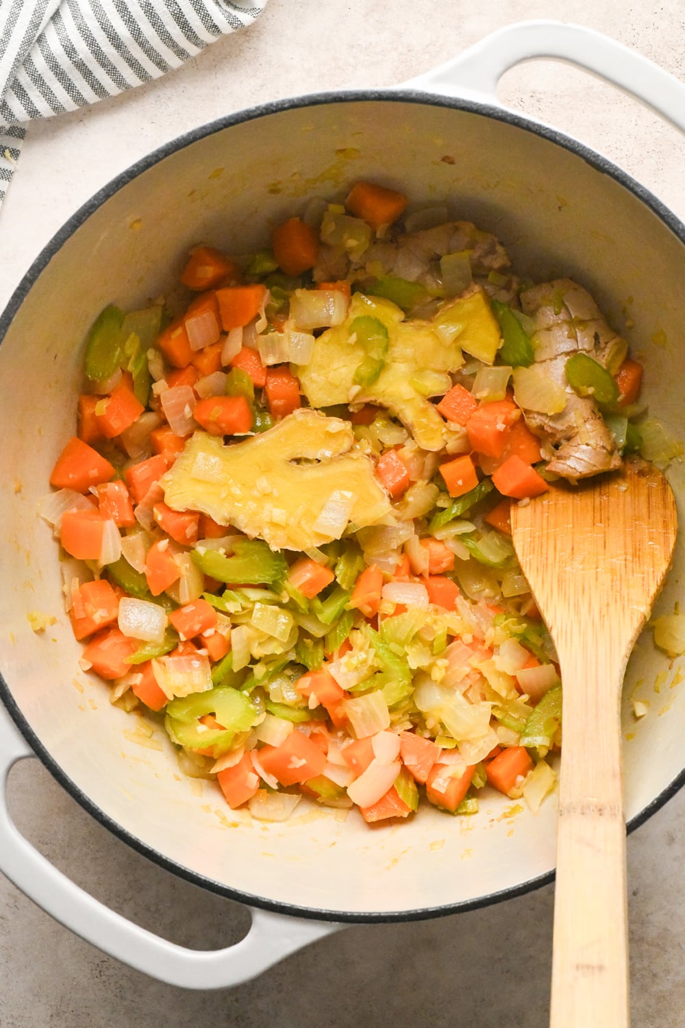 A white enameled pot containing a sautéed mixture of diced carrots, celery, and onions, with two large pieces of ginger, being stirred by a wooden spoon.