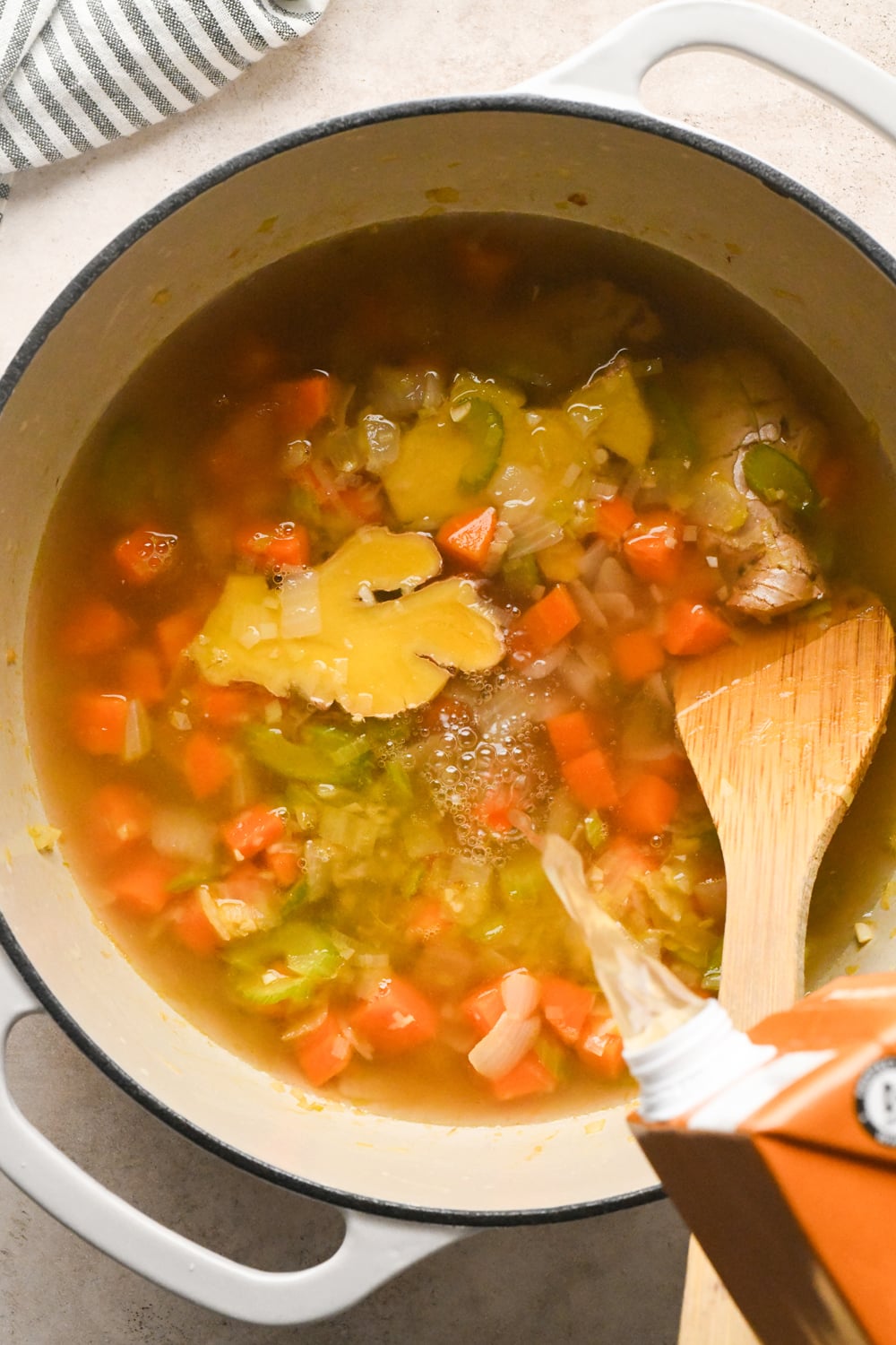 Broth being poured from a carton into a pot of vegetable soup with a wooden spoon and a slice of ginger.