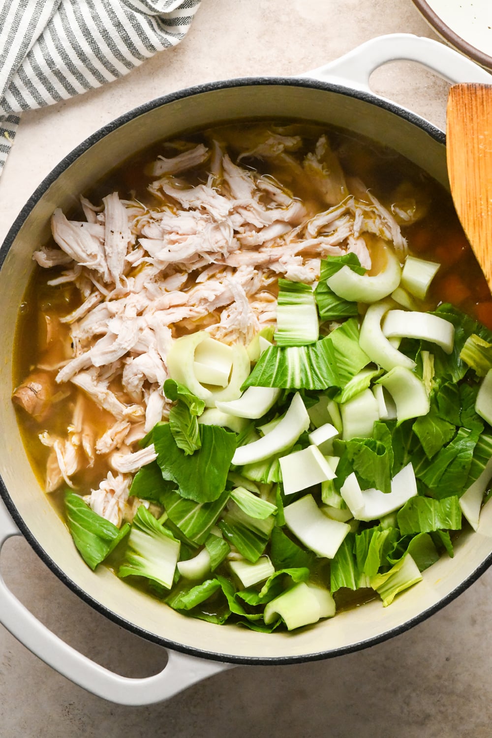 A white pot filled with broth containing shredded chicken and chopped bok choy.
