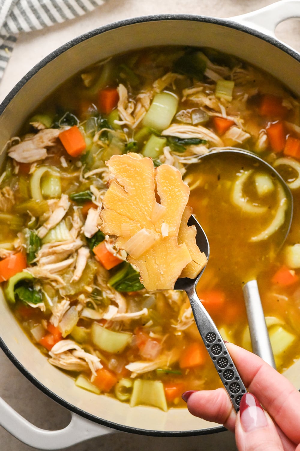 A spoon held over a pot of chicken vegetable soup, lifting a large, thick slice of ginger.
