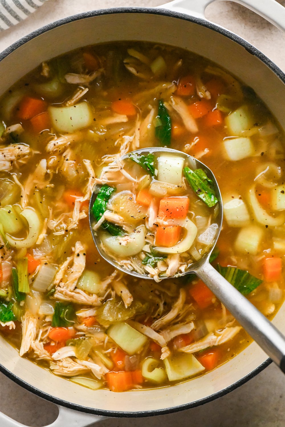 A ladle lifting chicken ginger soup with carrots, celery, and bok choy from a large pot.