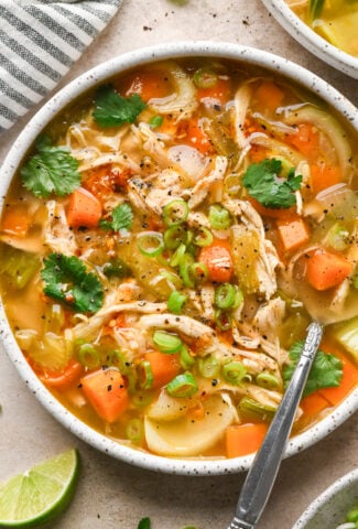 A bowl of ginger chicken soup with carrots, celery, green onions, and cilantro, served with a lime wedge.