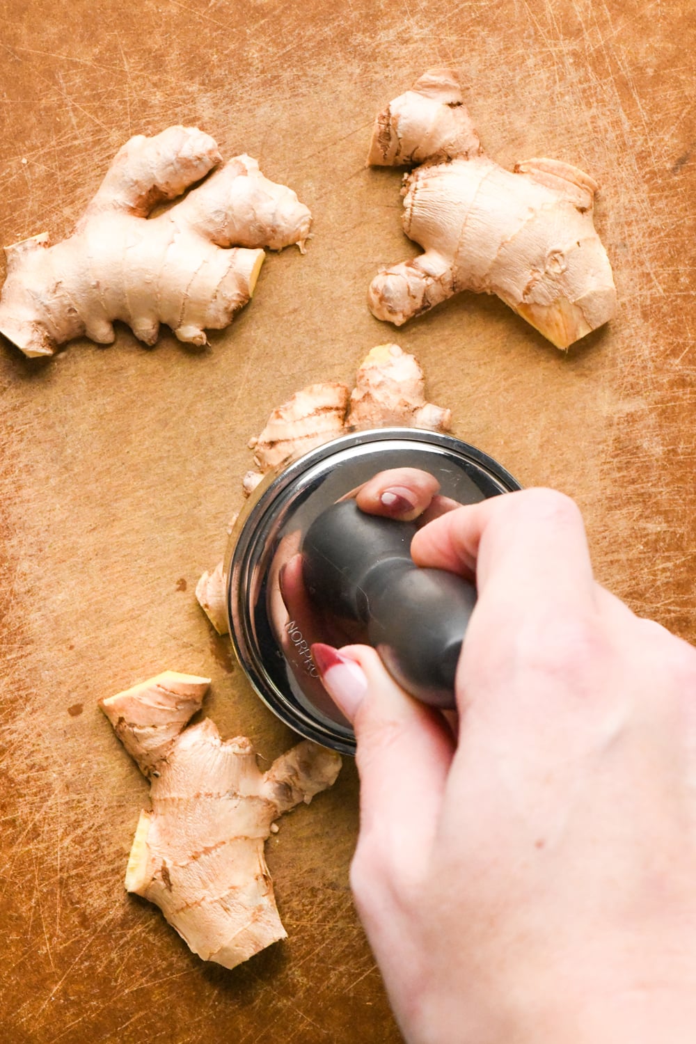 A hand uses a circular pounding tool to crush a piece of ginger on a wooden cutting board, with other ginger pieces nearby.
