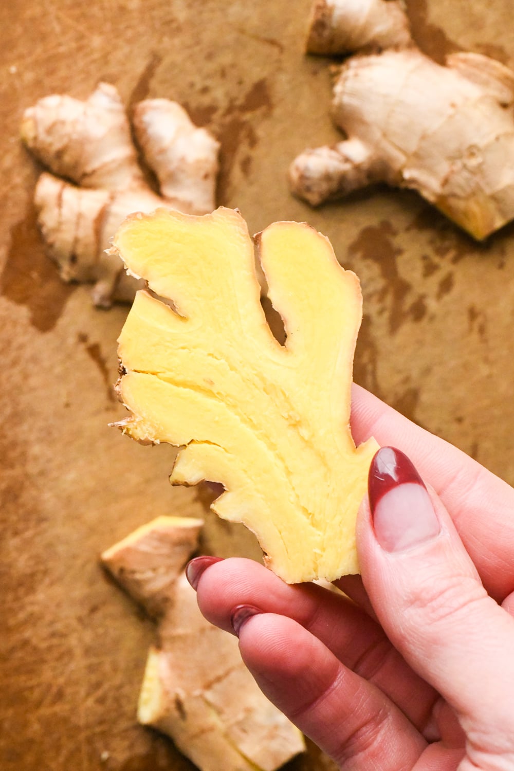 A hand holds a freshly sliced piece of bright yellow ginger root that has been smashed, with more ginger pieces resting on a wooden surface in the background.
