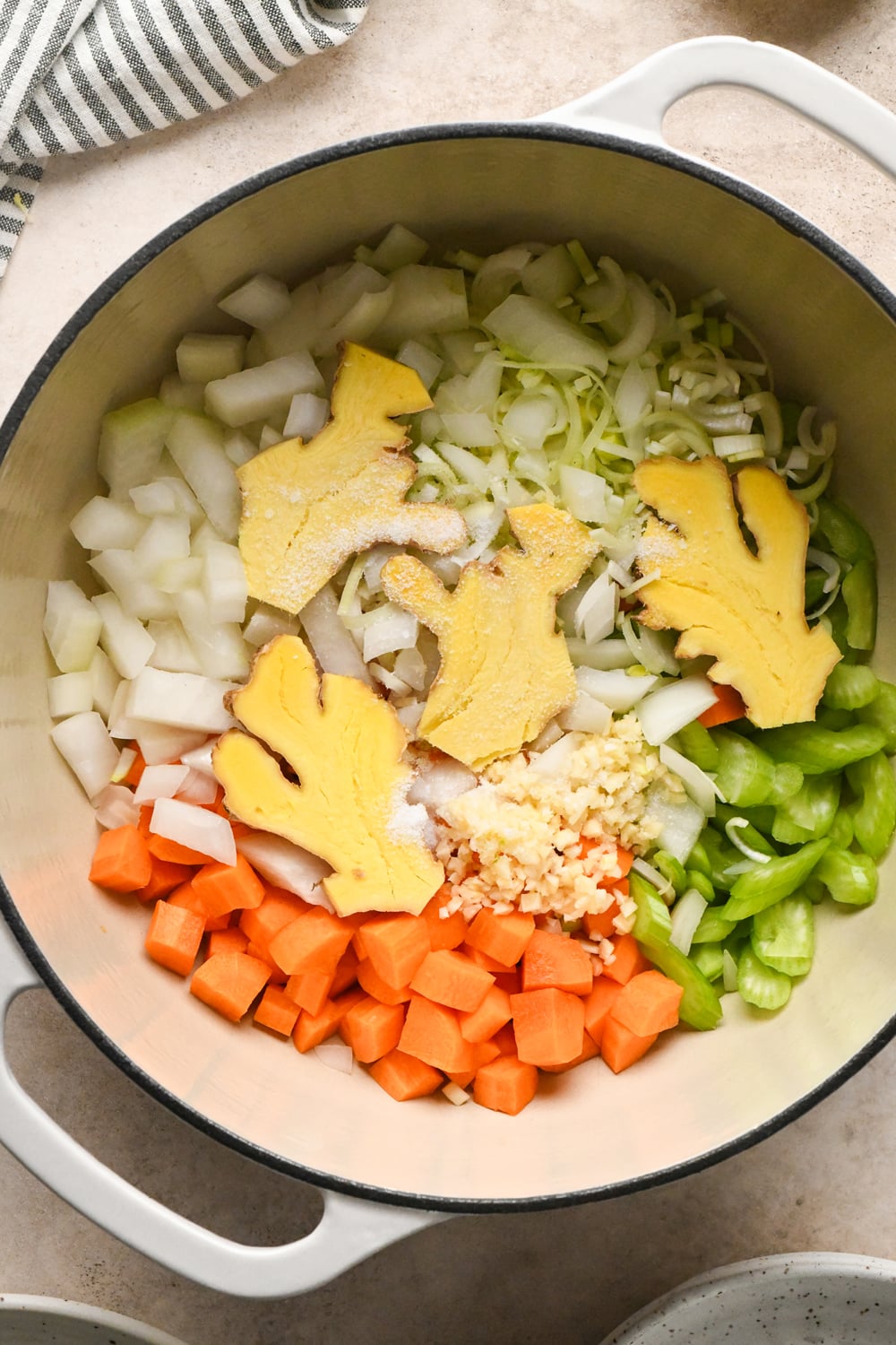 A white Dutch oven containing chopped onions, carrots, celery, minced garlic, and sliced fresh ginger.