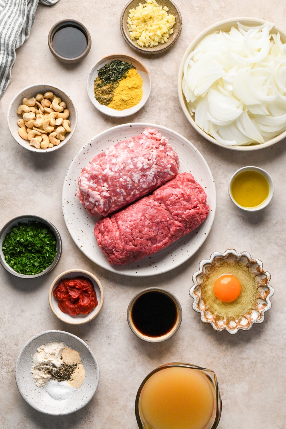An overhead view of ingredients for a smothered caramelized onion meatballs, including ground beef and pork,, sliced onions, raw cashews, an egg, chopped herbs, dried spices, and liquid condiments in various bowls.