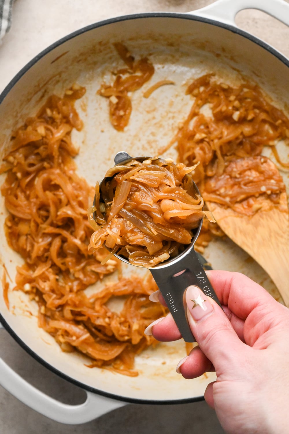 A hand holding a half-cup measuring scoop filled with golden-brown caramelized onions, held over a pan containing the rest of the onions.
